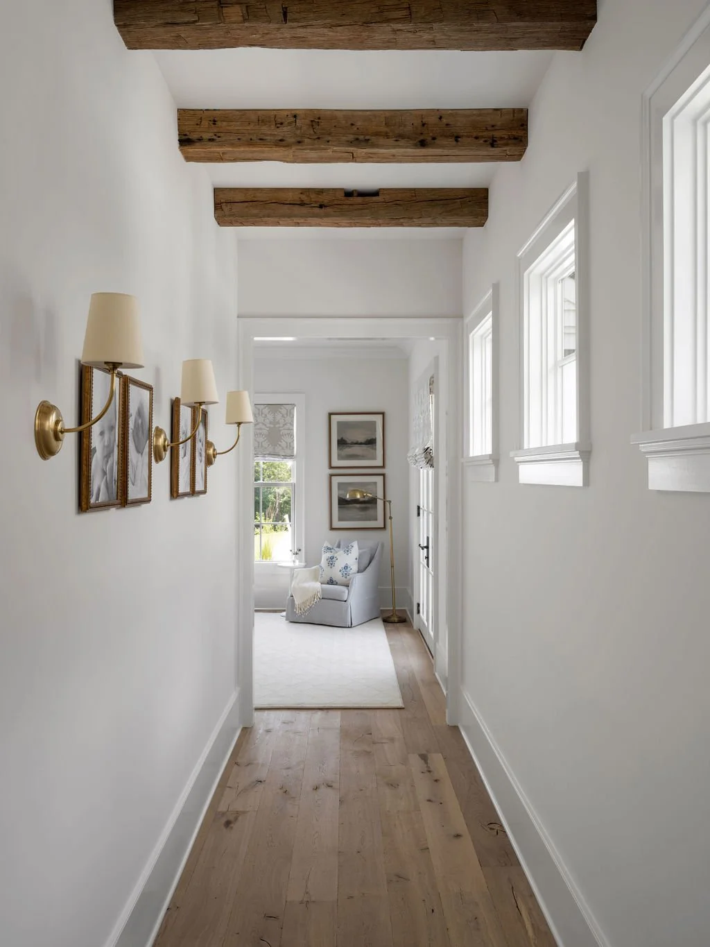 Bright hallway with wooden ceiling beams, white walls, and multiple windows, leading to a cozy sitting area with a white armchair and framed art.