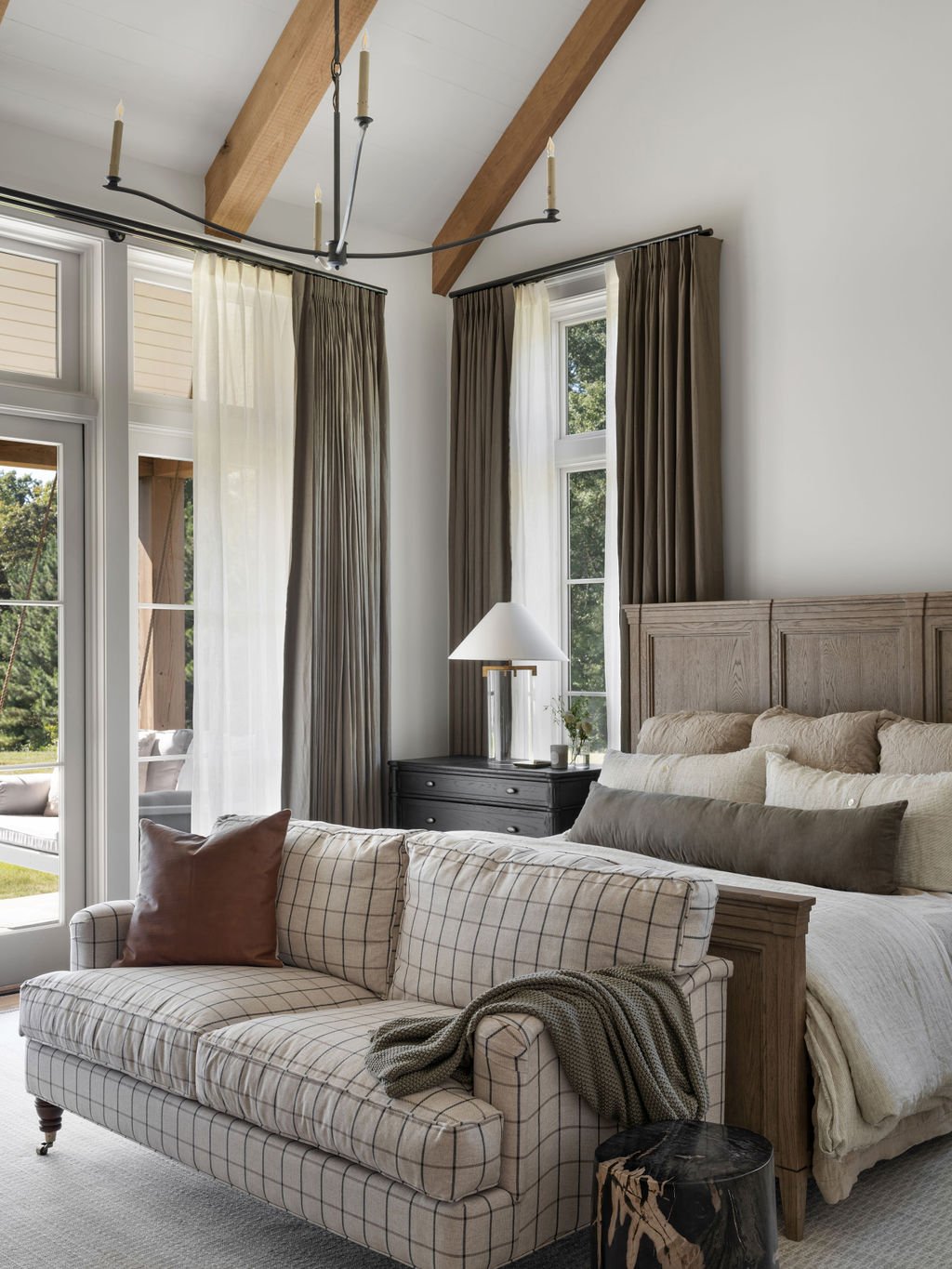 A bedroom with large windows, beige curtains, a wooden bed frame with beige bedding and multiple pillows, a dark gray nightstand with a white lamp, a plaid upholstered sofa with pillows, a tree trunk side table, and a ceiling with exposed wooden beam
