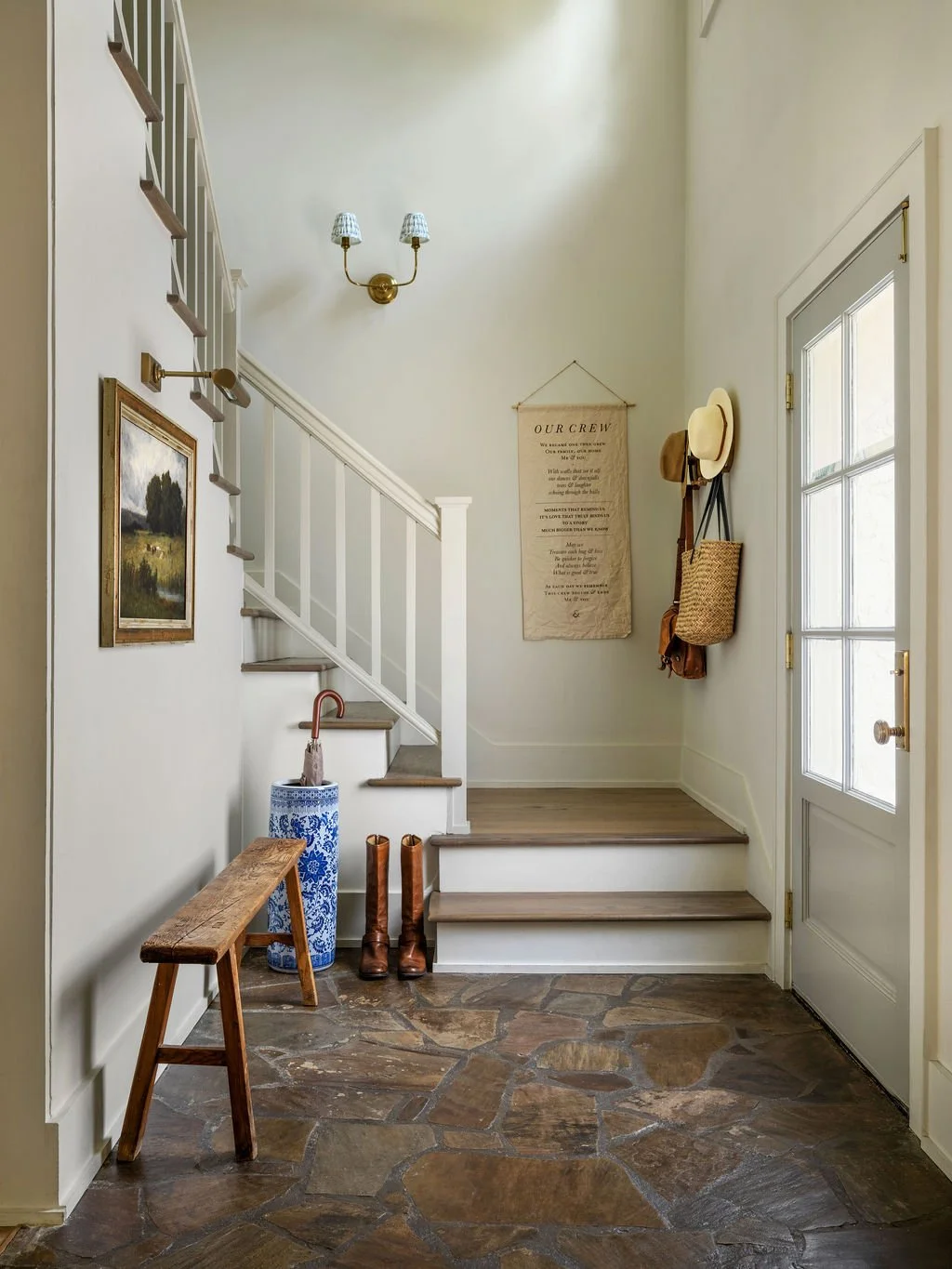 Entryway with stone floor, wooden bench, umbrella stand, books, shoes, and wall hooks holding hats and bags, near a door with glass panes.