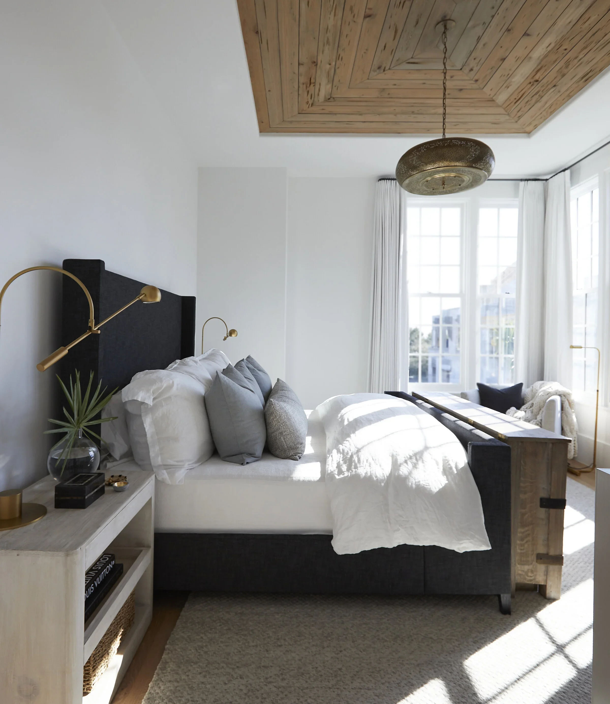 A bright bedroom with a white bed, gray and white pillows, a beige side table with a plant and books, large windows with white curtains, a wooden ceiling, and a gold circular pendant light.