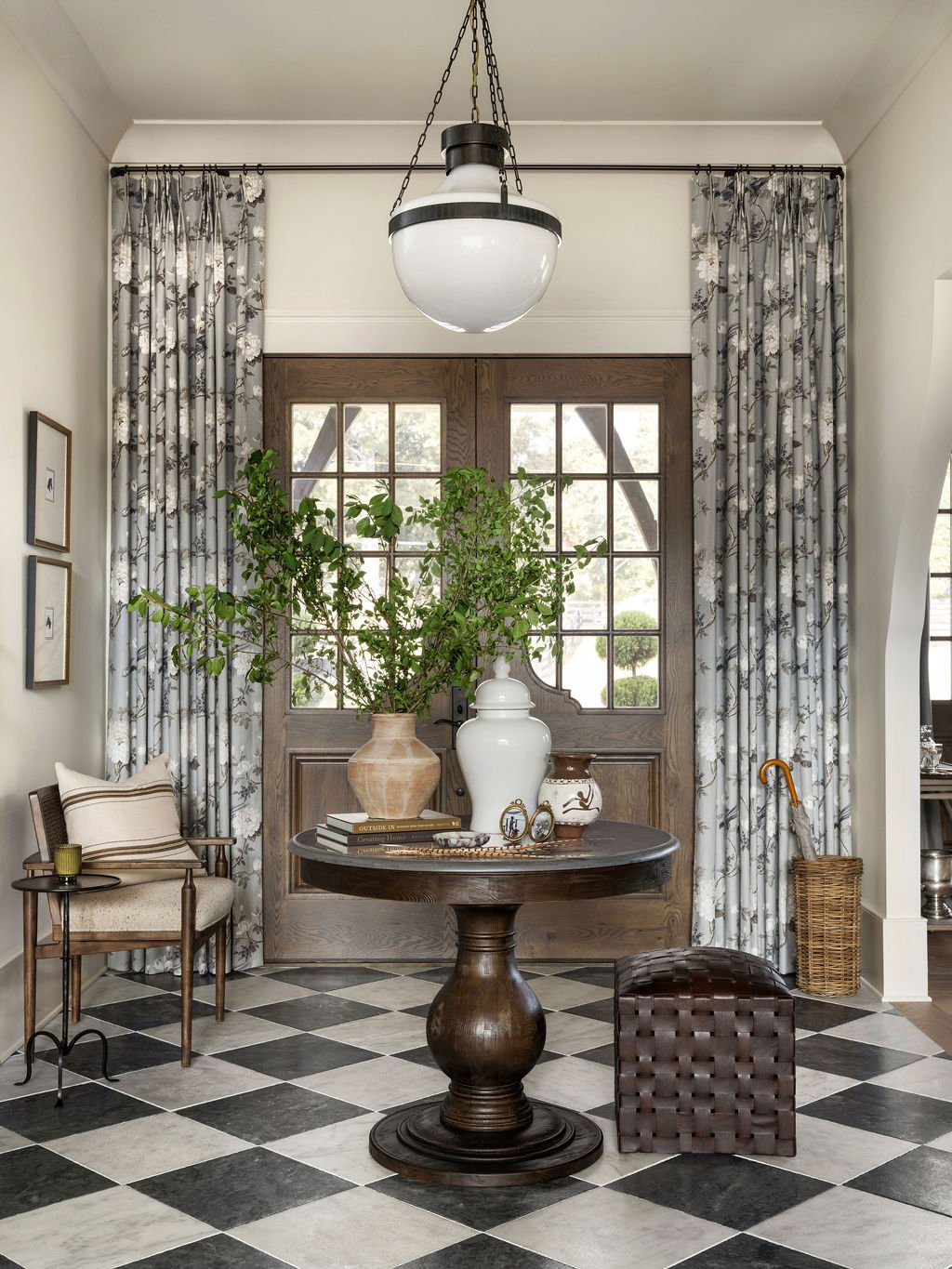 Living room with a round wooden table, large green plant in a tan vase, white decorative urn, framed pictures, checkered black and white floor, large window with floral curtains, a bench with a pillow, umbrella stand, and a wicker basket.