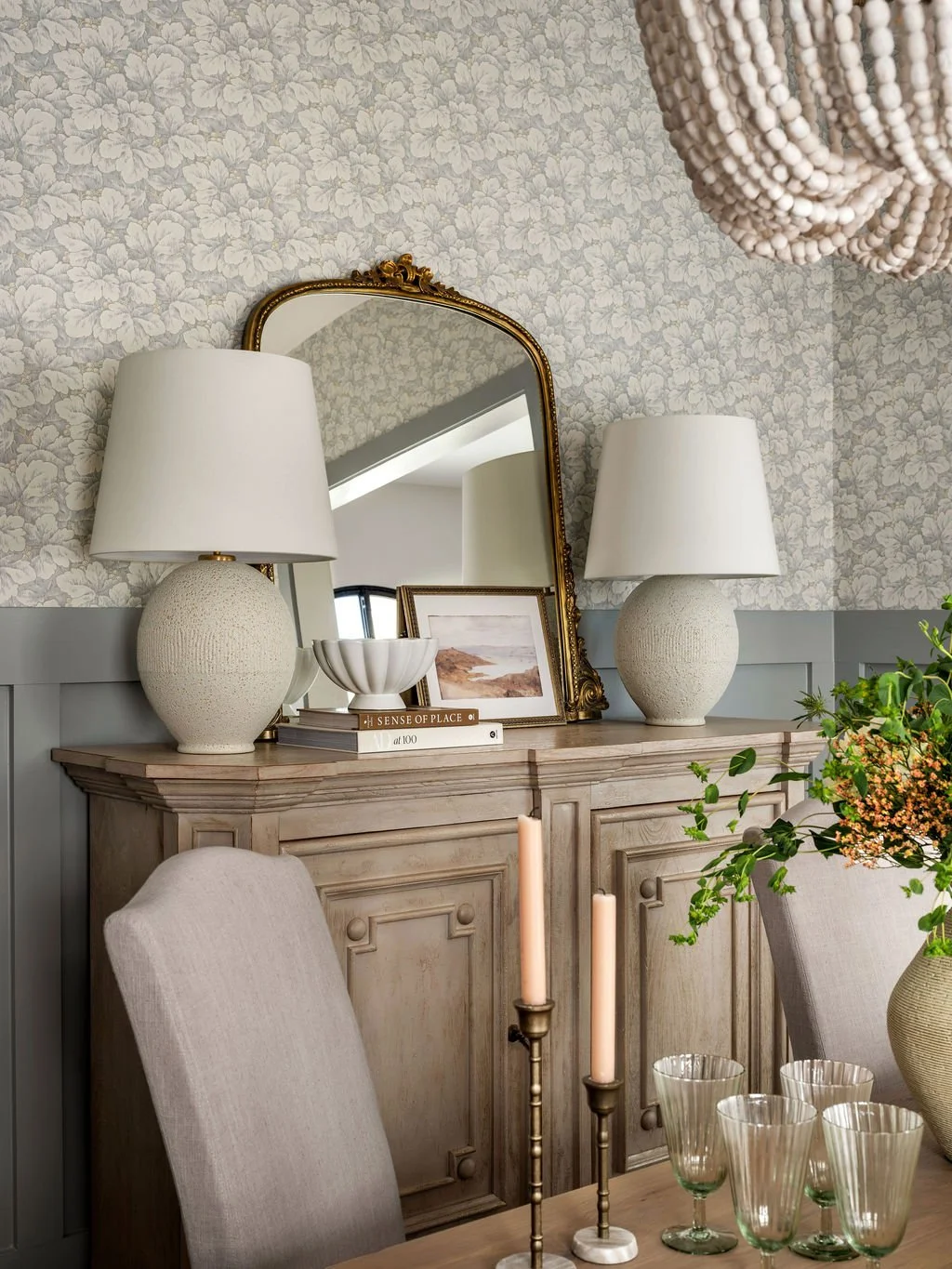 A wooden sideboard with two large white lamps, framed artwork, a mirror, books, and a bowl on top. In front, a dining table with beige upholstered chairs, a pair of candlesticks with tall pink candles, and a vase with flowers. The room has floral wal
