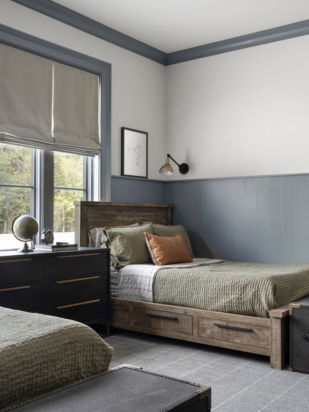 Bedroom with a wooden bed frame, green bedding, pillows, a black dresser with a globe, a window with a gray Roman shade, and wall decor including a framed sketch and a wall-mounted light.