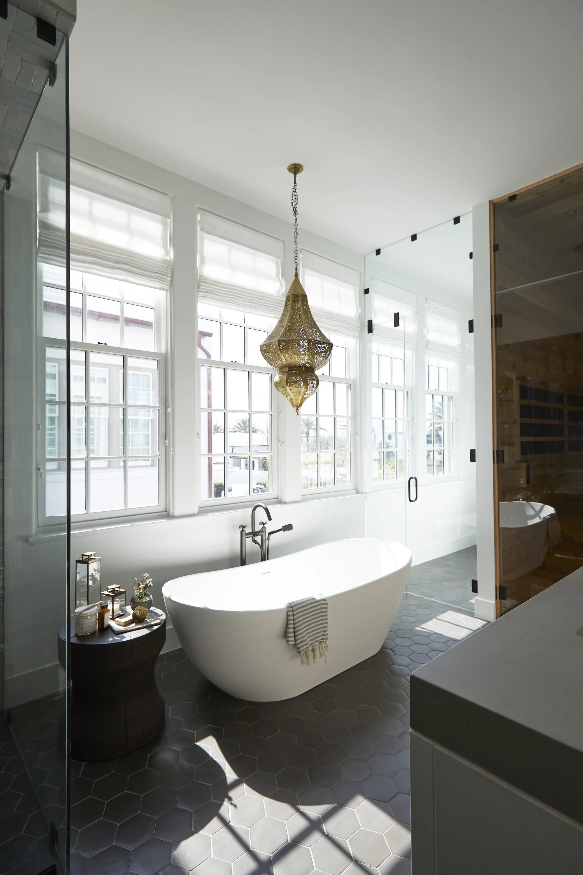 Bright bathroom with a freestanding bathtub, large windows, a hanging brass chandelier, a side table with decor, and a glass shower enclosure.