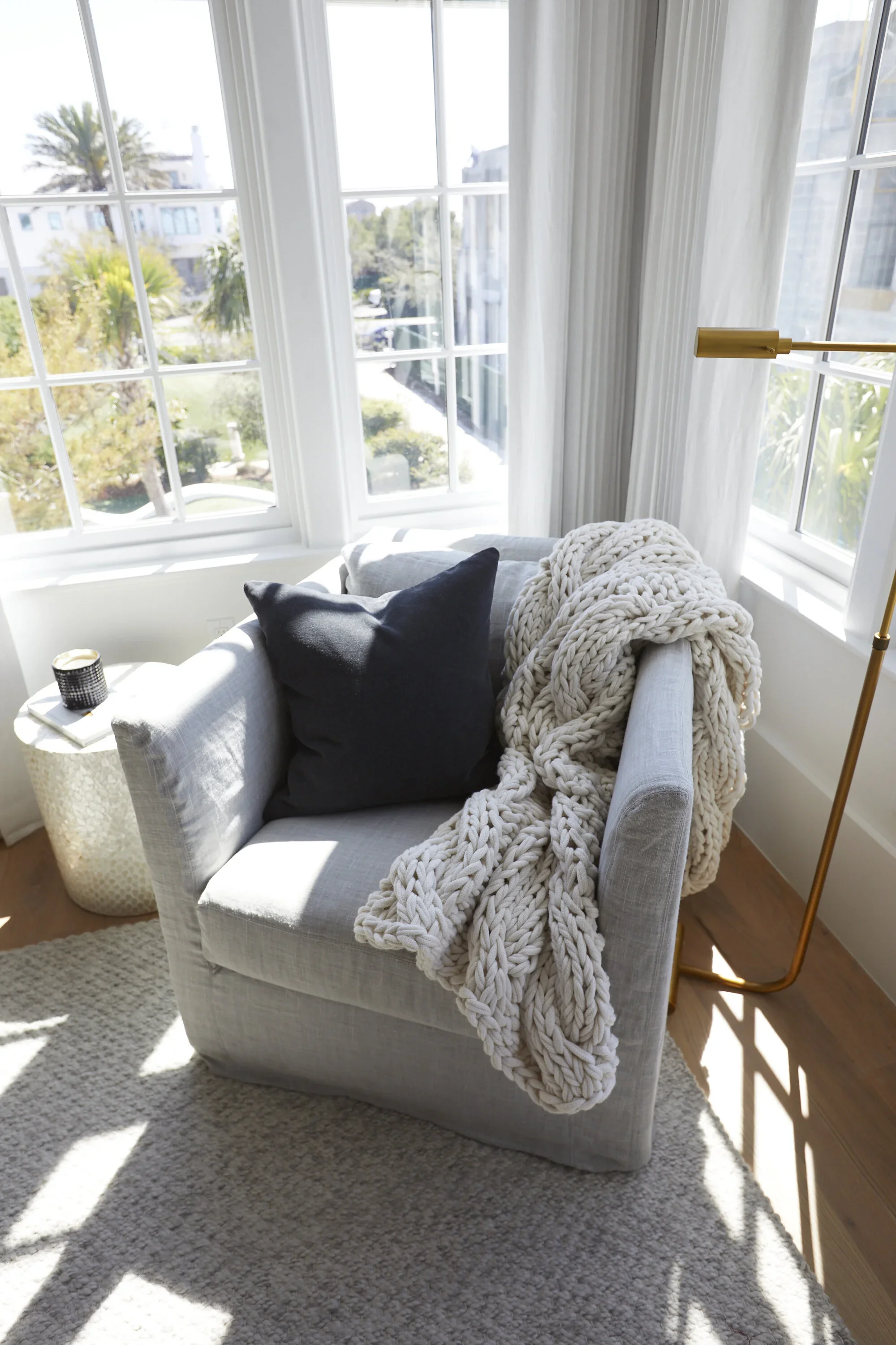 A cozy corner with a white armchair, black pillow, chunky knitted blanket, beside a small side table with a candle, and surrounded by large windows with sheer curtains showing a sunny outdoor scene with palm trees.
