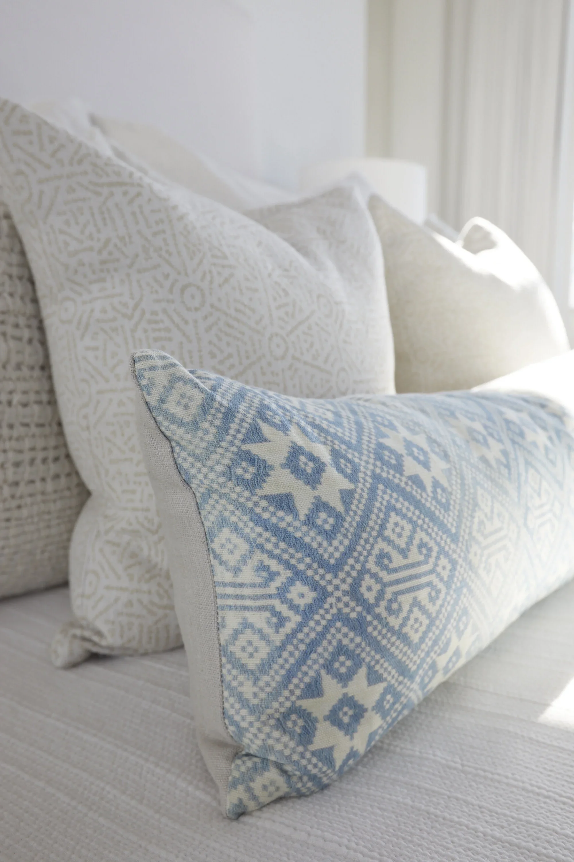 Close-up of decorative pillows on a bed, including a rectangular pillow with blue and white geometric patterns, and other white pillows with textured fabric, illuminated by sunlight.