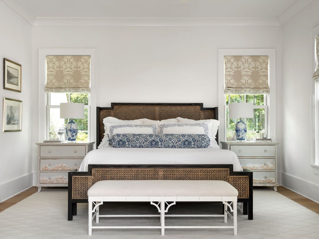 A bedroom with a large bed, white bedding, and decorative pillows, flanked by two nightstands with lamps, and a white bench at the foot of the bed, in a bright room with windows and artwork on the walls.