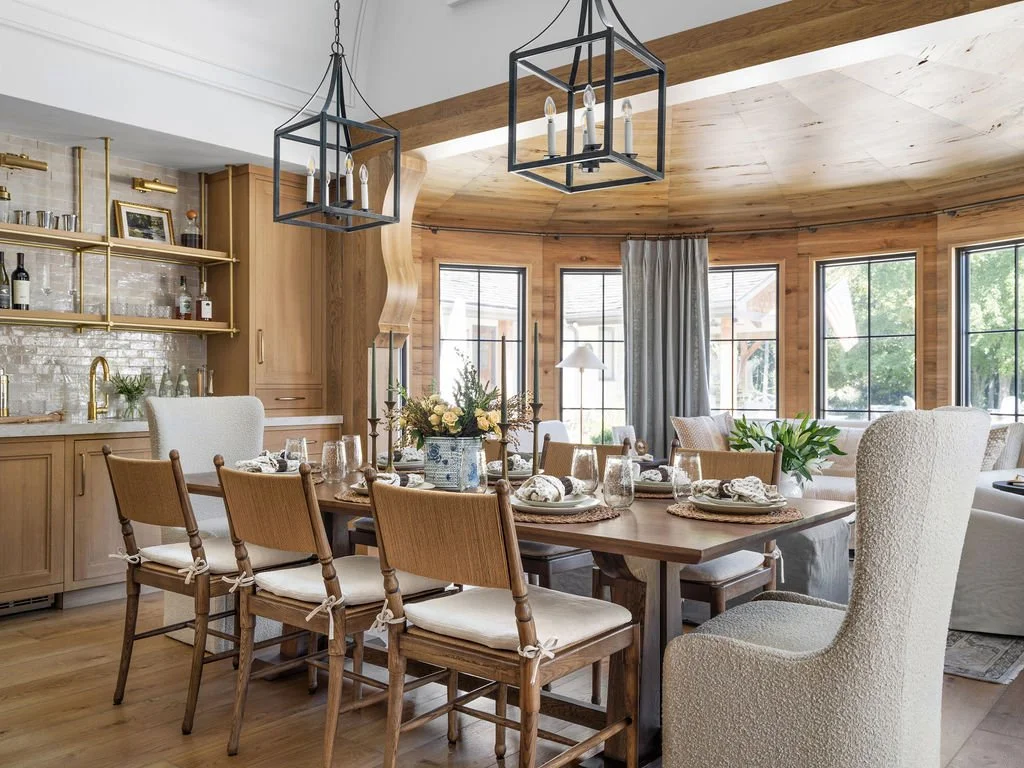 A dining area with a large wooden table set with plates, glasses, and napkins, surrounded by wooden chairs and two white armchairs, in a room with large windows, wooden flooring, and a vaulted wooden ceiling. Overhead, two black geometric chandelier 