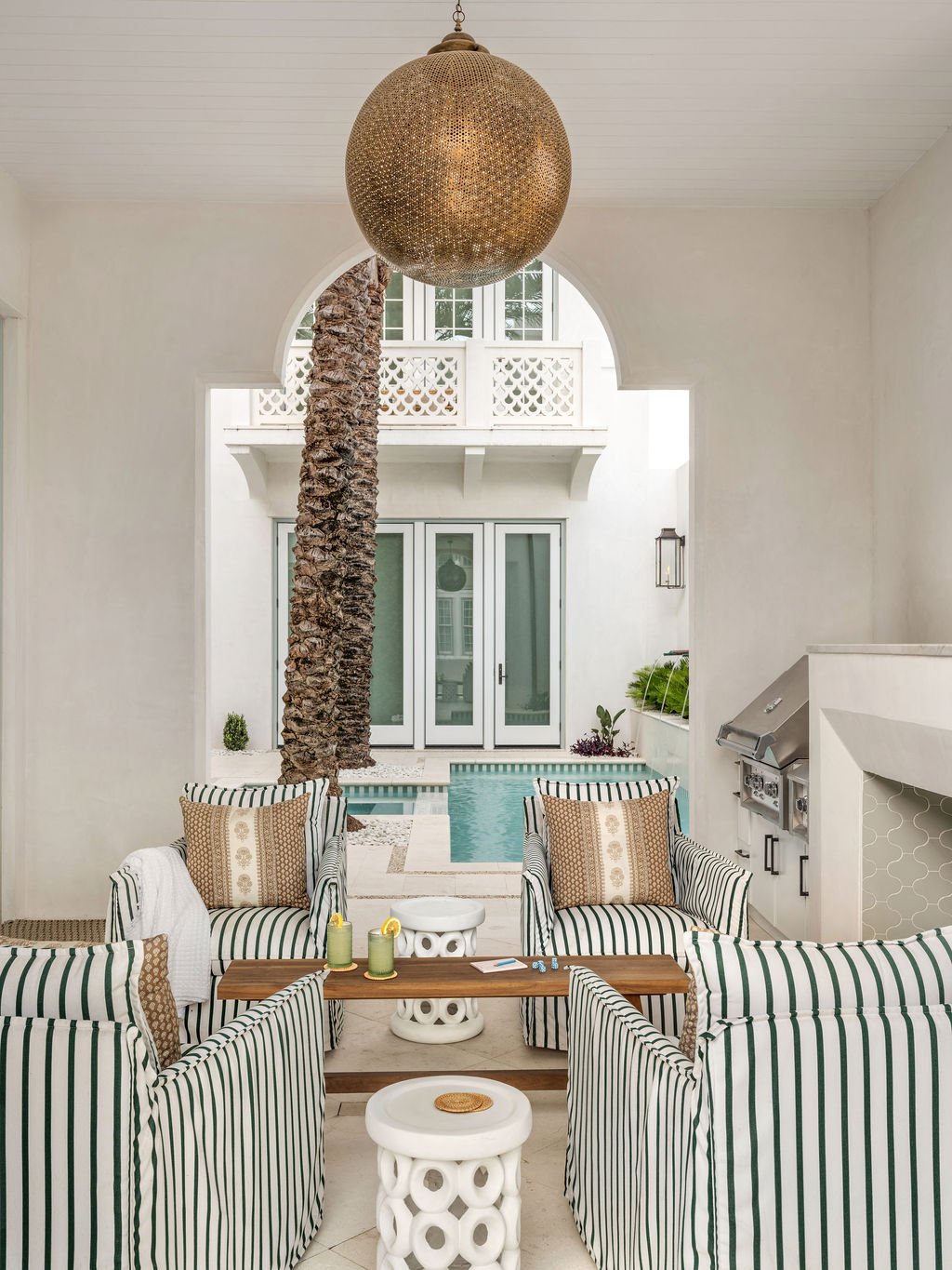 Indoor outdoor living space with striped armchairs, a palm tree, and a pool visible through an archway, under a large hanging light fixture.