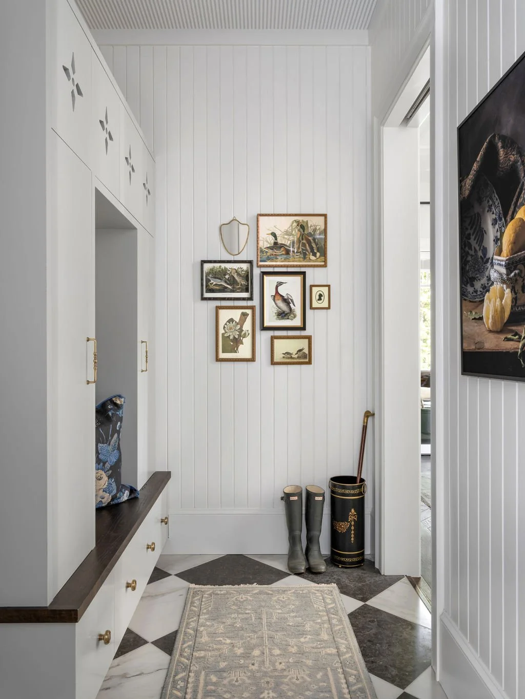 Entryway with white beadboard walls, framed artwork of birds and nature, black and gold umbrella stand, black rain boots, and a small bench with a decorative pillow.