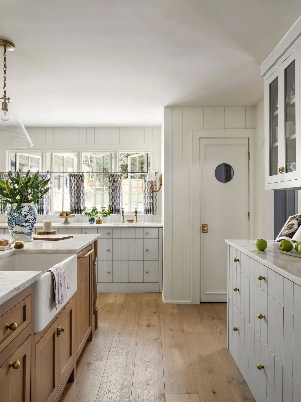 A bright kitchen with white wood paneling, a large window with patterned curtains, a kitchen sink with a vase of green foliage, and wooden cabinets with round brass knobs. There are green apples on the counter and a book open nearby.