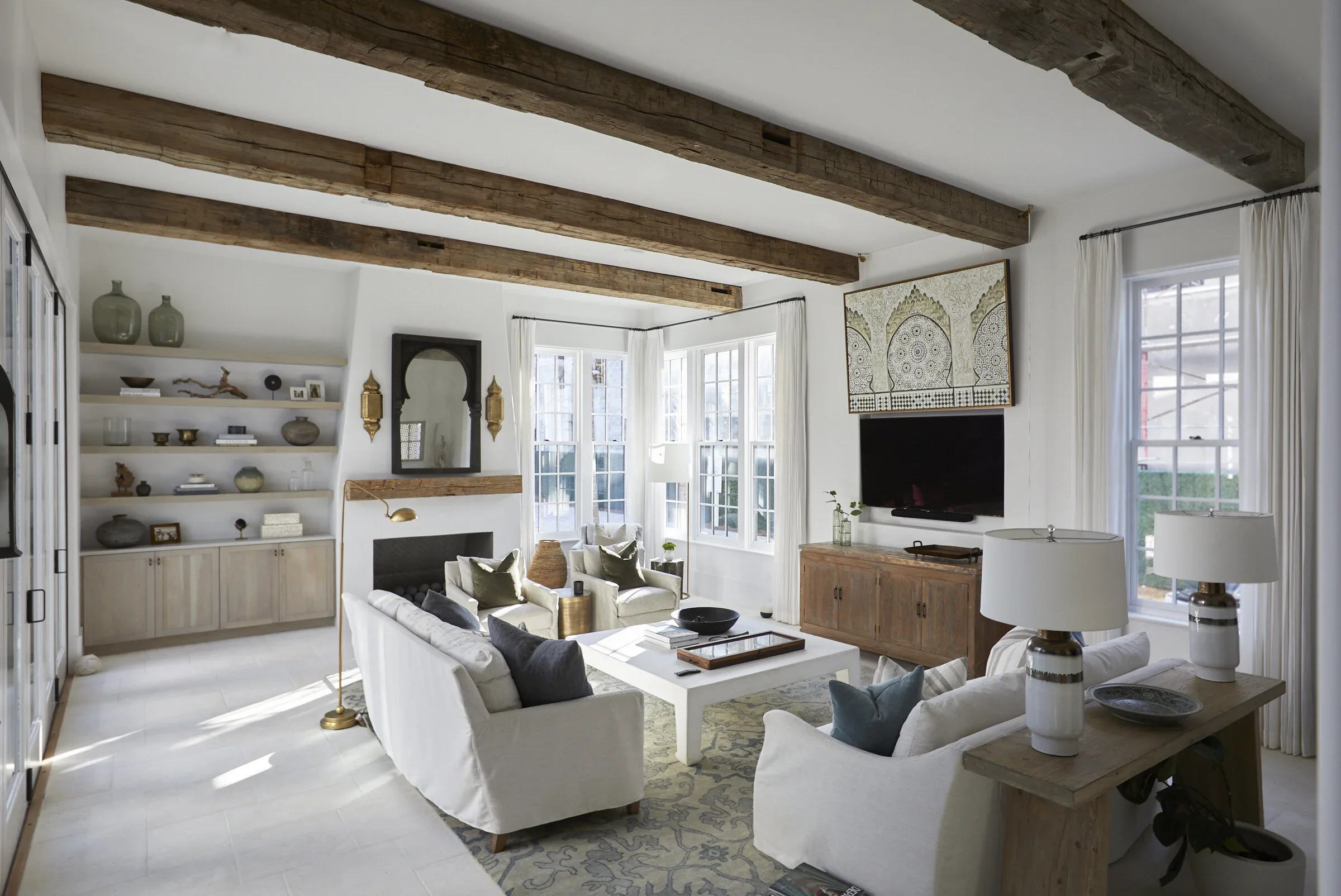 Bright living room with white couches and armchairs, a white coffee table, wooden ceiling beams, large windows with white curtains, built-in shelves with decor, a fireplace, and a mounted flat-screen TV.
