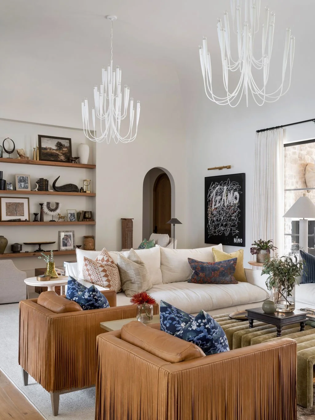 Living room with white sofas, colorful pillows, two tan leather armchairs, a wooden-interior bookshelf, modern black and white art, and large chandeliers in a bright space.