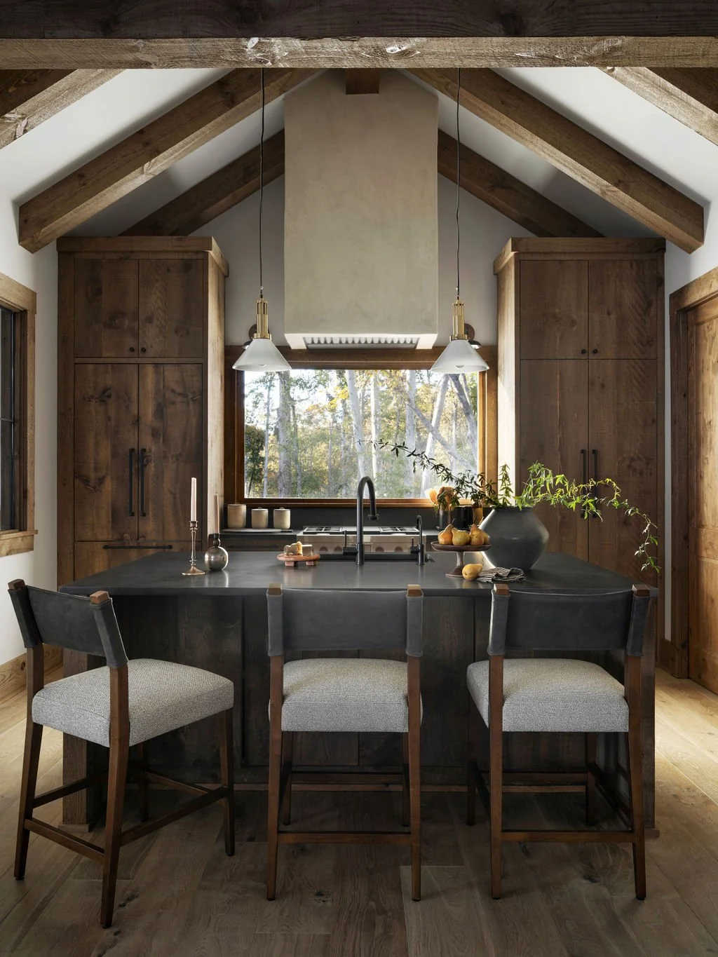 A modern kitchen with wooden cabinets, a black kitchen island with bar stools, large window overlooking trees, and pendant lights hanging from the wooden vaulted ceiling.