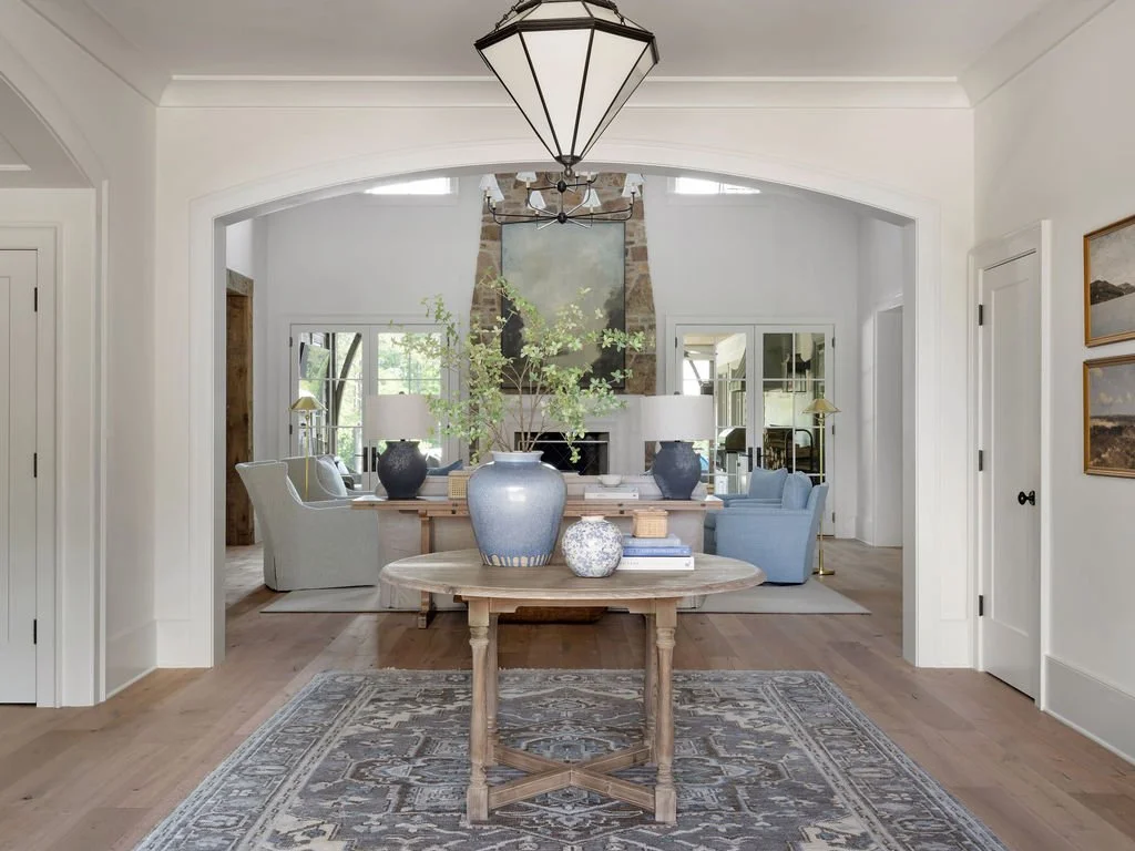A spacious, elegant living room with a high ceiling, large fireplace, and a mix of traditional and modern furnishings. Central table with decorative vases and greenery. Soft natural light fills the room.