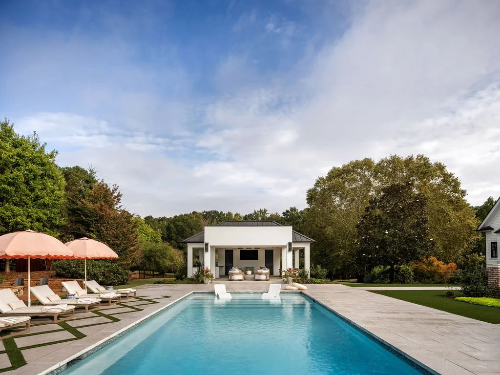 Resort-style backyard pool with lounge chairs and umbrellas, surrounded by greenery and a modern pool house.