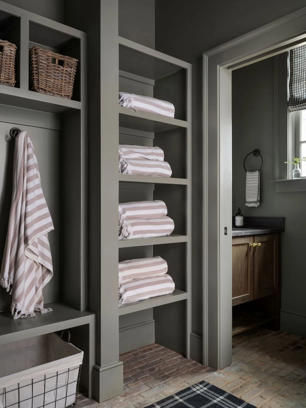A linen closet with shelves holding folded striped towels, next to a hanging towel, in a bathroom with a wooden vanity and a window.