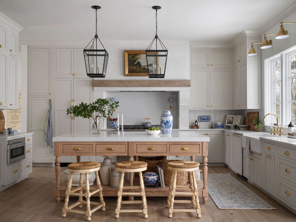 Bright, white kitchen with a centered island table, four wooden stools, and a windowed corner. Decor includes plants, blue and white ceramics, and gold cabinet hardware.