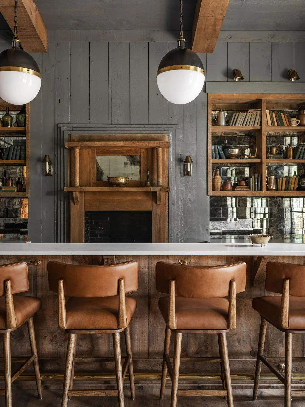 A cozy kitchen interior with a wooden fireplace, open shelving filled with books and decorative items, pendant lights, and leather bar stools at a white countertop.