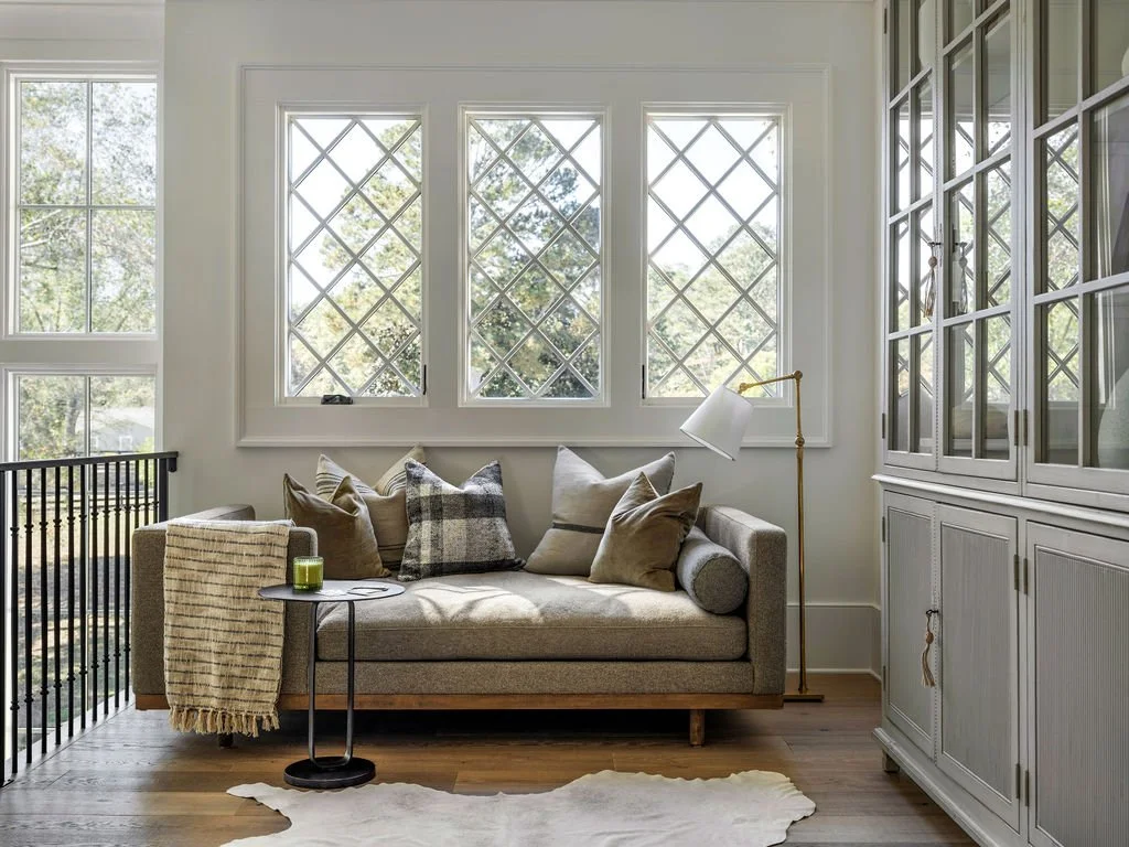 Living room with a gray sofa, decorative pillows, a side table with a candle, a floor lamp, large windows with a view of trees outside, and a white cabinet on the right.