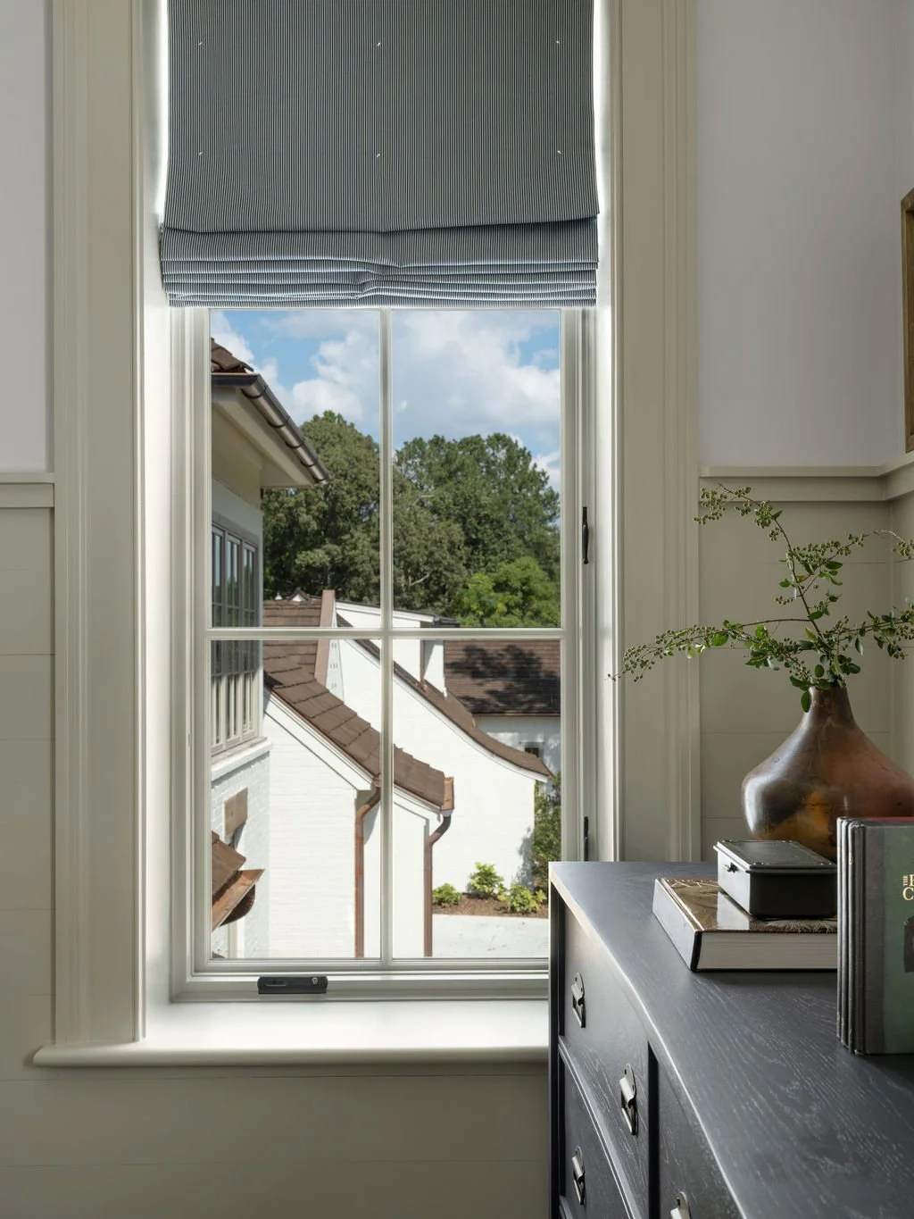 View through a window showing a neighboring house with white walls and brown roof, surrounded by green trees and a partly cloudy sky.