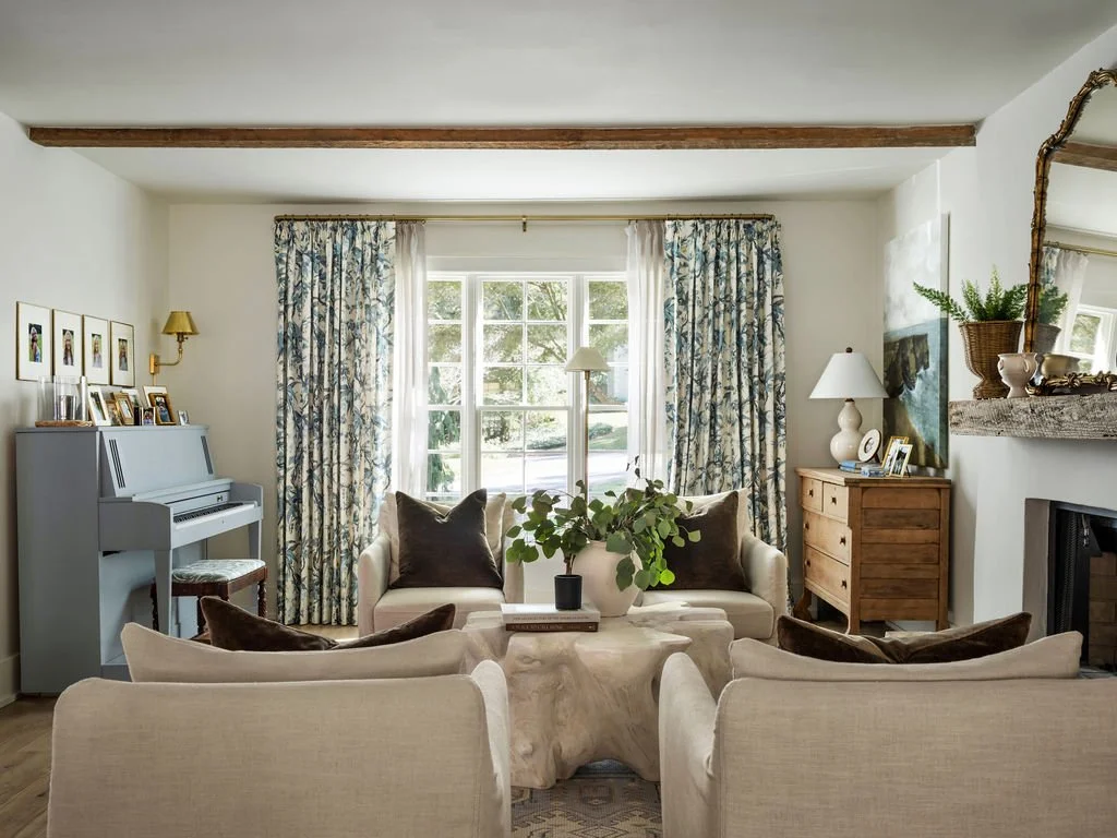Bright living room with cream sofas, a central coffee table with a large plant, floral curtains, a white piano on the left, and a wooden cabinet with lamps and framed photos on the right. A large window with trees outside.