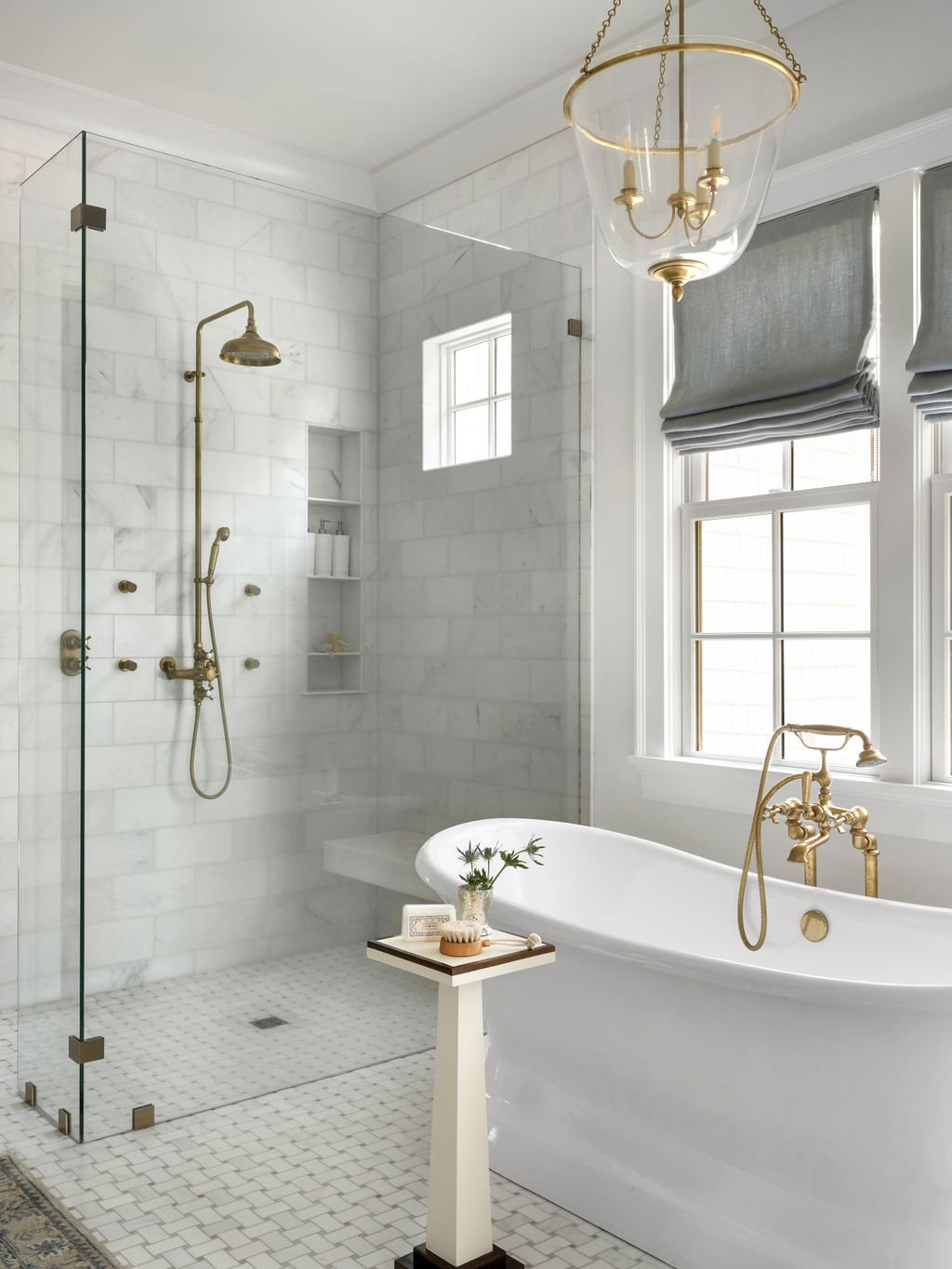 A luxurious bathroom featuring a white marble walk-in shower with gold fixtures, a freestanding white bathtub with gold fixtures, and two large windows with gray Roman shades. There is a small white side table with a plant, soap, and a brush, and a c