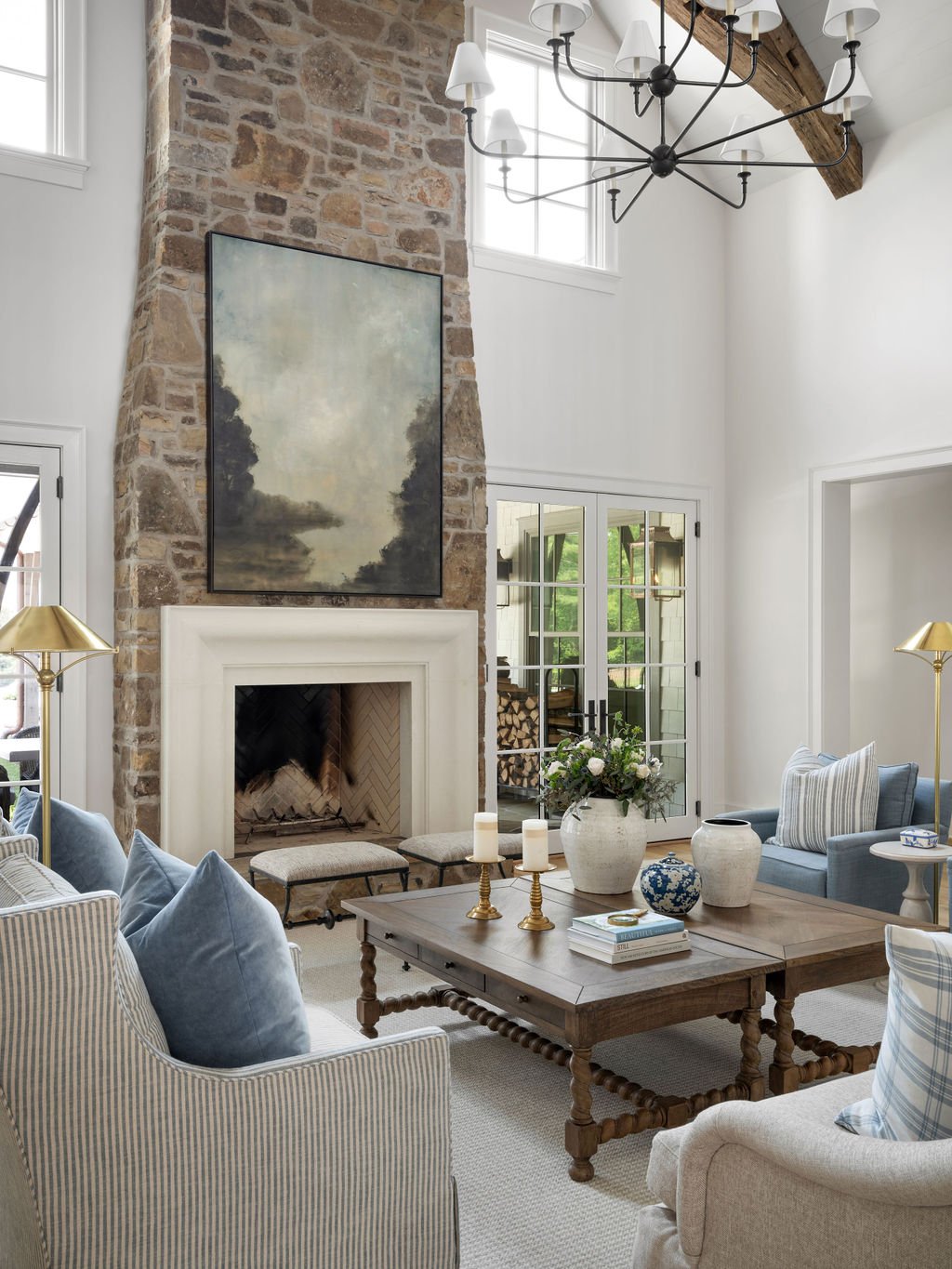 A living room with a tall brick fireplace, a modern chandelier, large windows, and glass doors leading outside. The room is decorated with sofas, pillows, a wooden coffee table with vases and candles, and a view of a firewood storage area outside.