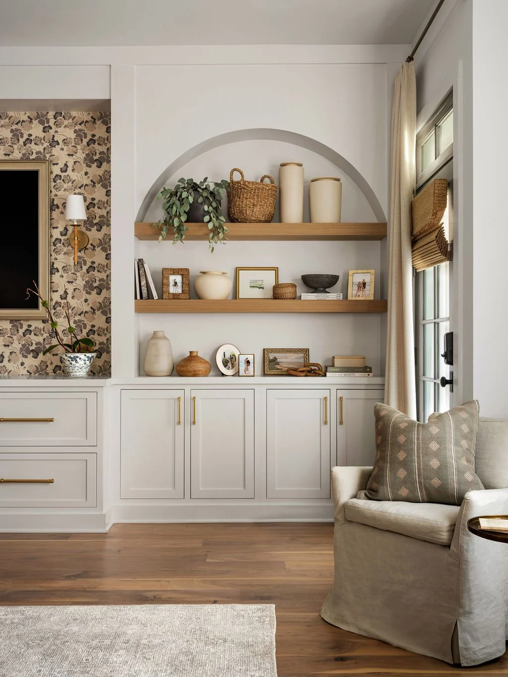 Decorative living room with built-in shelving, displaying vases, framed photos, and small sculptures, with a beige armchair and a window with curtains on the right side.