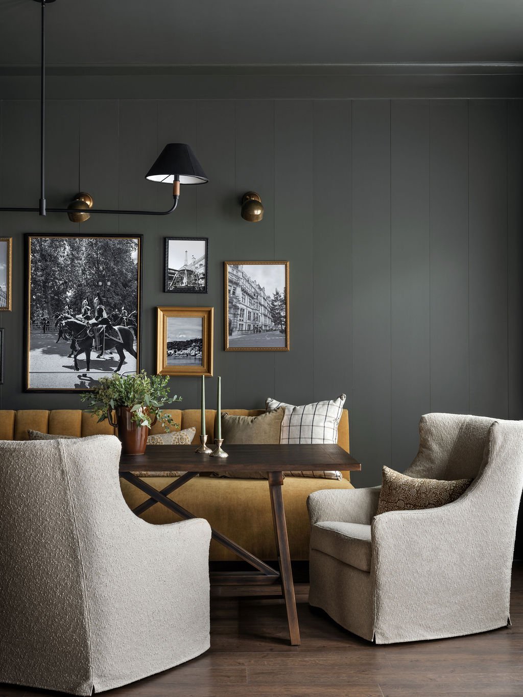 A cozy living room with a dark green wall decorated with framed black and white photographs, a mustard-colored sofa, two off-white armchairs, a wooden table with a potted plant and two tall candlesticks, and a modern black wall-mounted lamp.