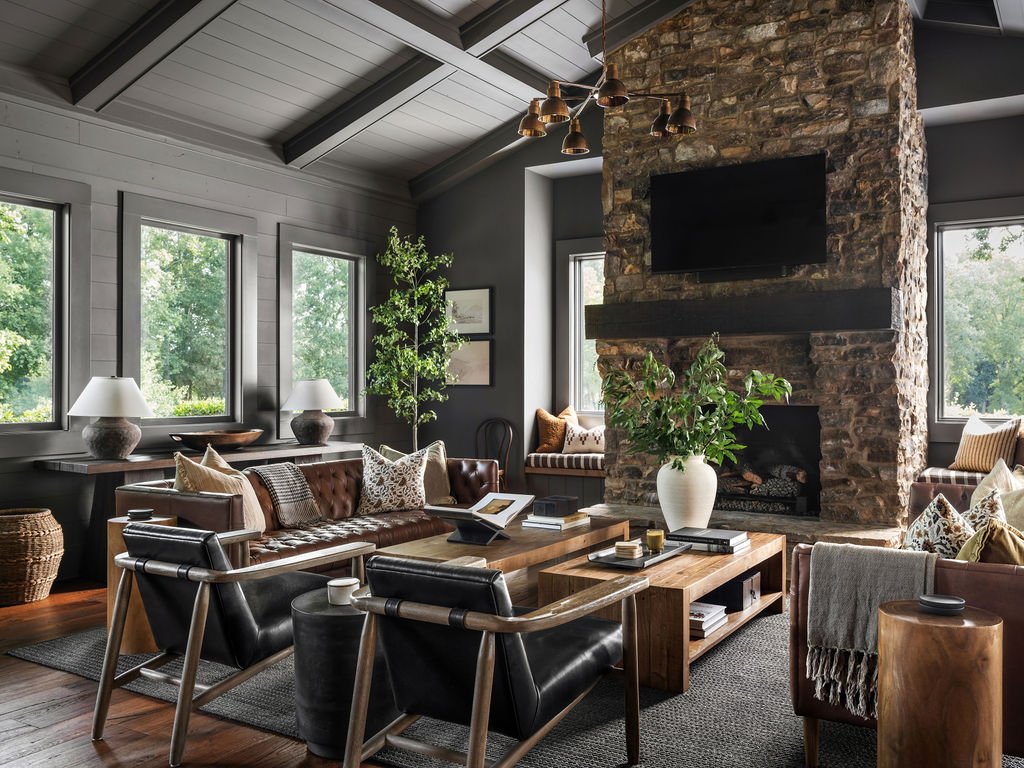 Living room with a stone fireplace, large windows, leather and fabric furniture, wooden coffee table, and potted plant.