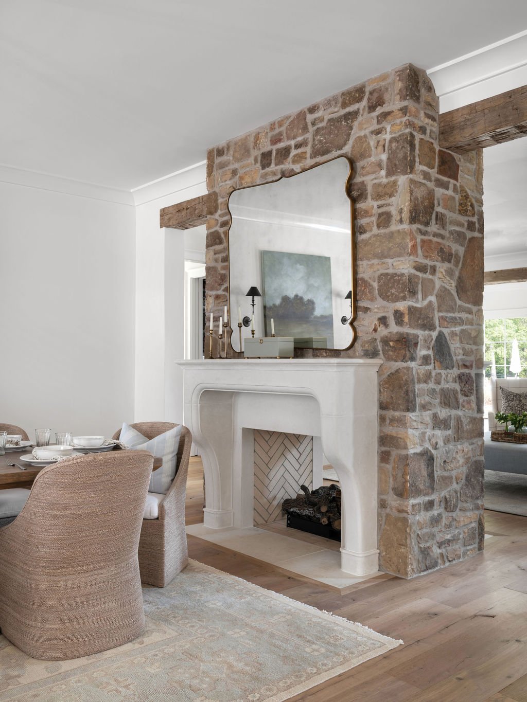 Interior of a living room with a stone fireplace, white mantel, and a large mirror above it. Part of a dining table with chairs is visible on the left. There are decorative items on the mantel and a painting reflected in the mirror.