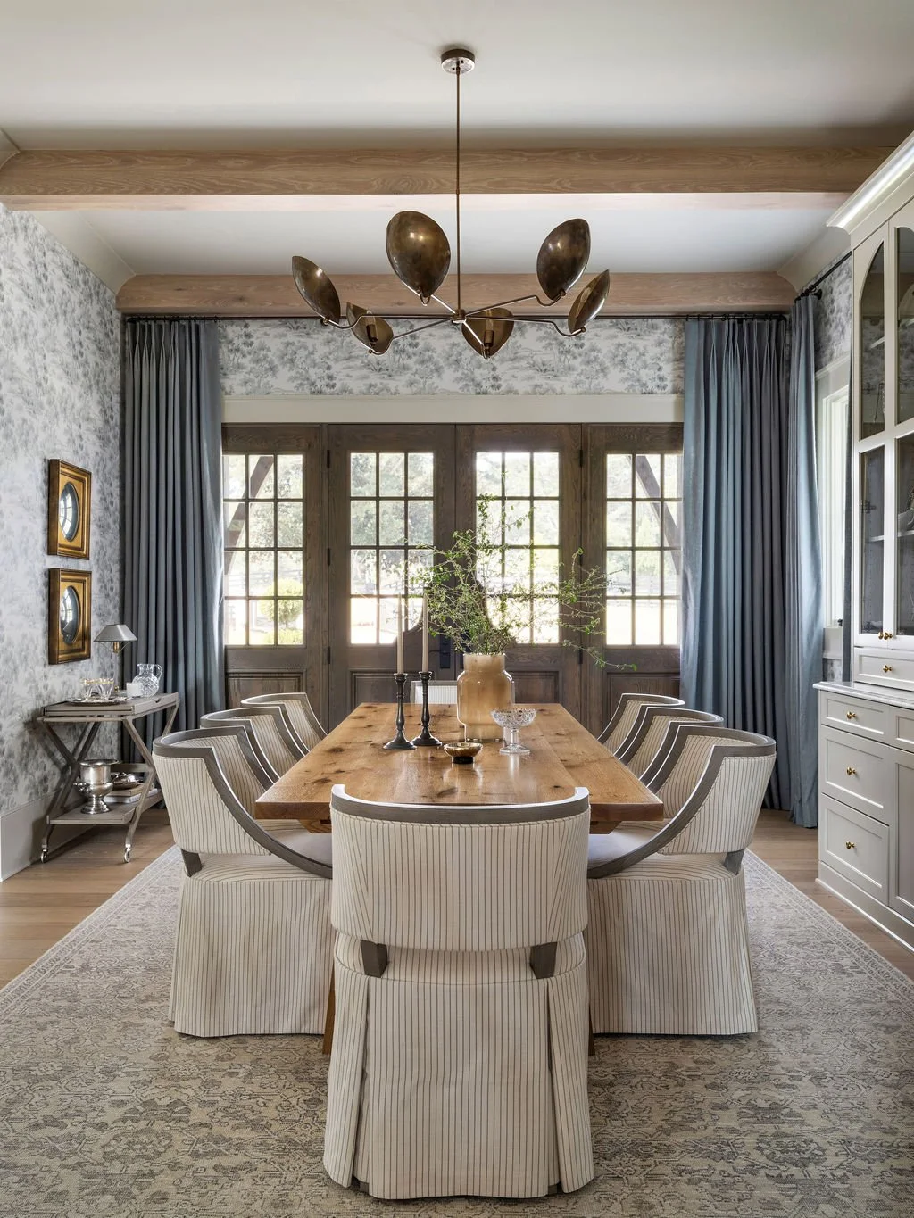 A rustic dining room with a wooden table, eight upholstered chairs, and decorated with a large ceramic vase with greenery, candlesticks, and glass bowls. The room features wood-paneled double doors, patterned wallpaper, gray curtains, and a modern chandelier.