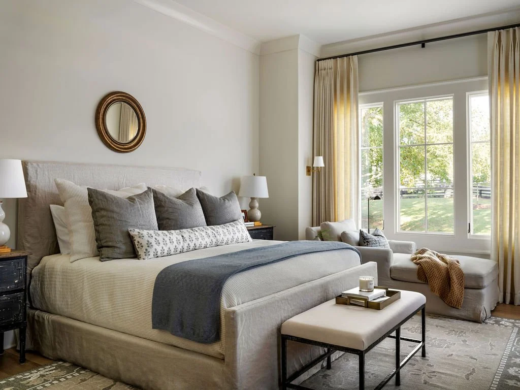 A bedroom with a large bed, gray and white pillows, a gray blanket, a cushioned bench at the foot, and a window with beige curtains showing a green outdoor scene.