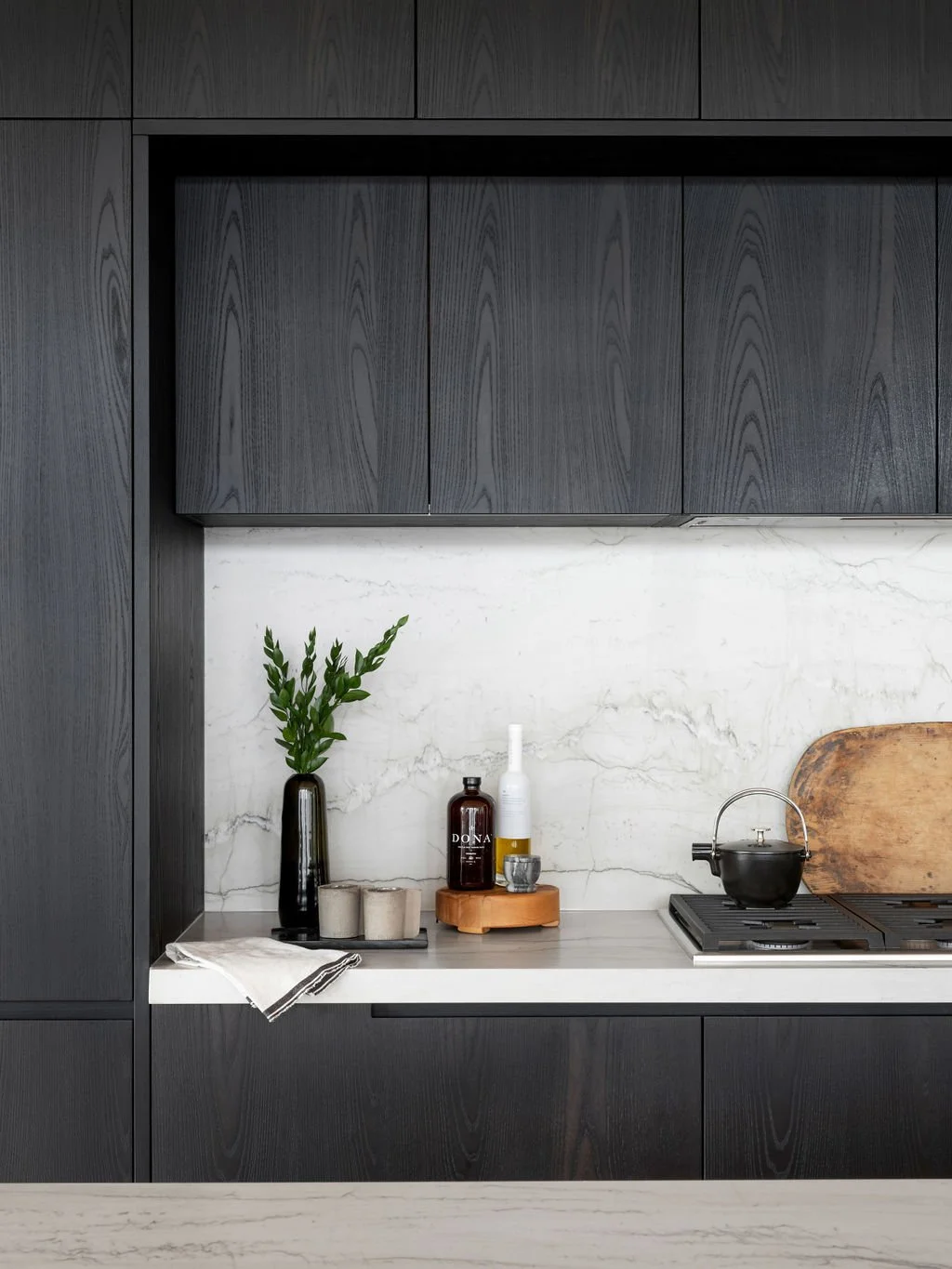 Modern kitchen with dark wood cabinets, marble backsplash, green plant in a black vase, bottles, and a black teapot on a stove.