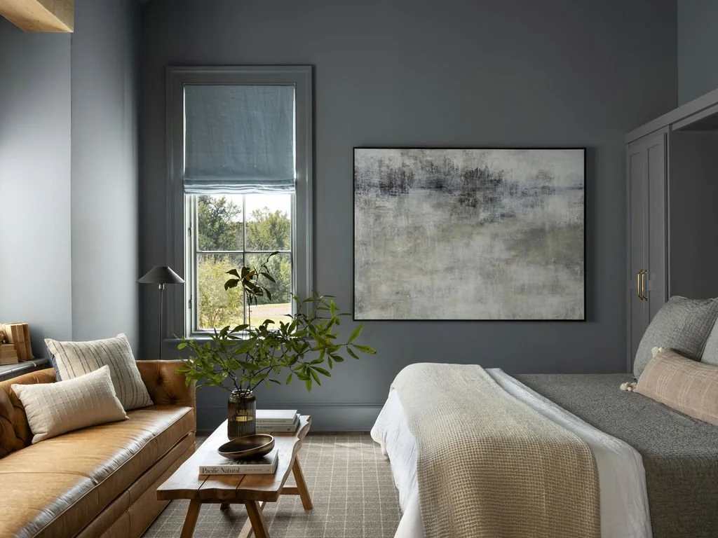 A modern bedroom with blue-gray walls, a window with a blue shade, a bed with gray and beige bedding, a gray wardrobe, a side table, a plant, and an abstract painting.