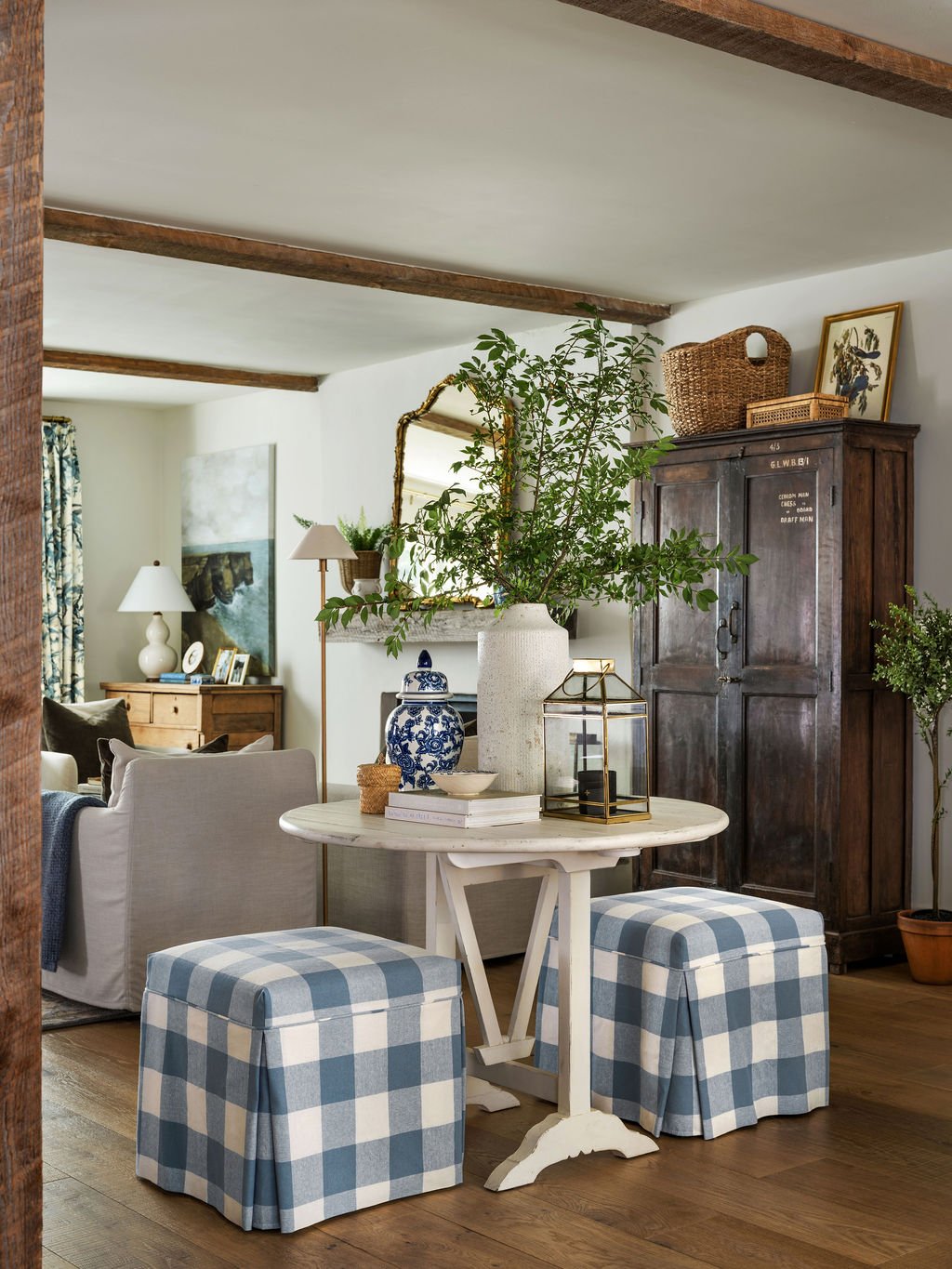A cozy living room and dining area with a white round table, two blue and white checkered ottomans, a large vase with green branches, a blue and white porcelain jar, and a lantern. There is a dark wood cabinet, framed artwork, and a mirror on the wal