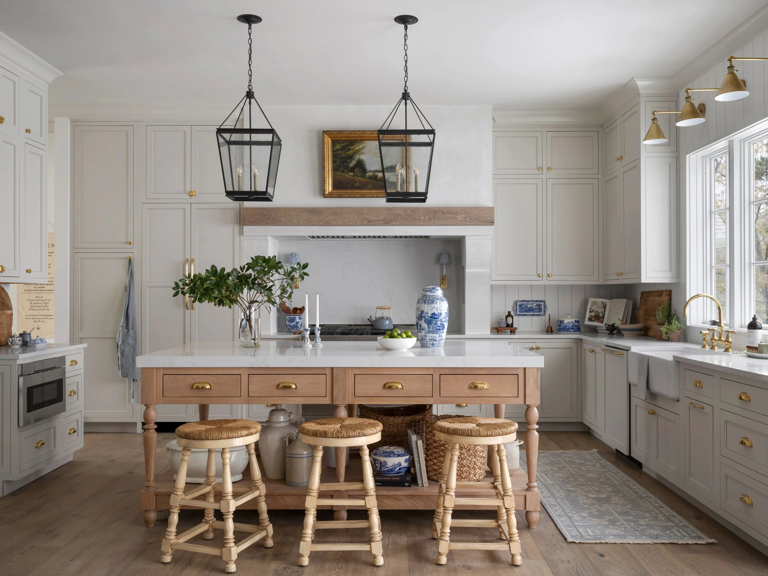 Bright kitchen with white cabinets, a central island with a wooden base, and stools. Decor includes plants, blue and white ceramics, and gold hardware. Large windows and pendant lights are featured.