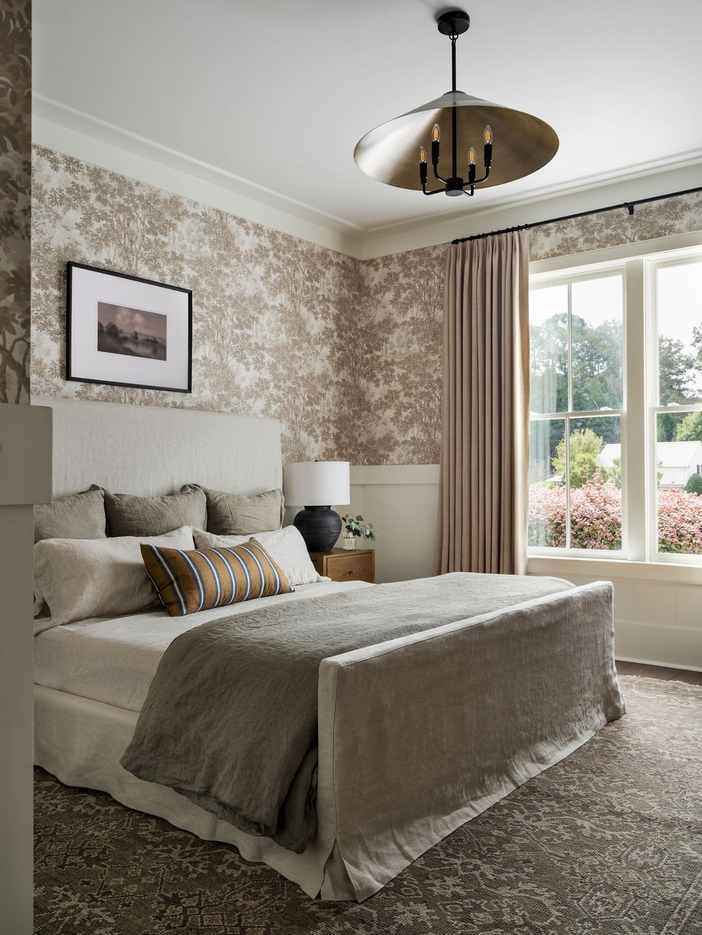 A neatly made bed with beige and brown bedding and pillows in a bedroom with floral wallpaper, a window with beige curtains, a nightstand with a lamp, and a modern chandelier.