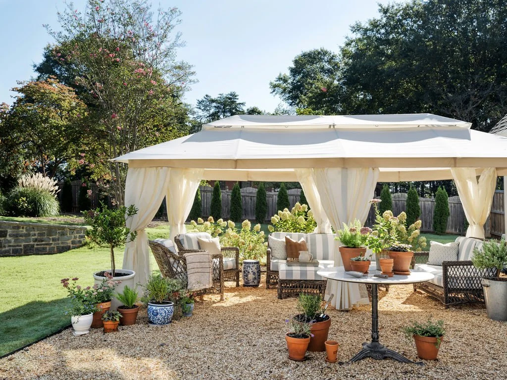 A backyard patio with a white canopy tent, outdoor seating, and potted plants on a gravel surface surrounded by green trees and a wooden fence.