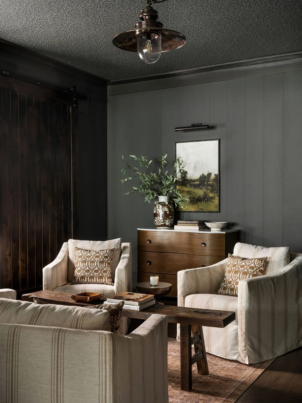 A cozy living room with beige striped armchairs, a rustic wooden coffee table, a dark wooden sideboard, and a large green vase with plants. The walls are gray with a landscape painting, and there's a vintage ceiling light fixture.