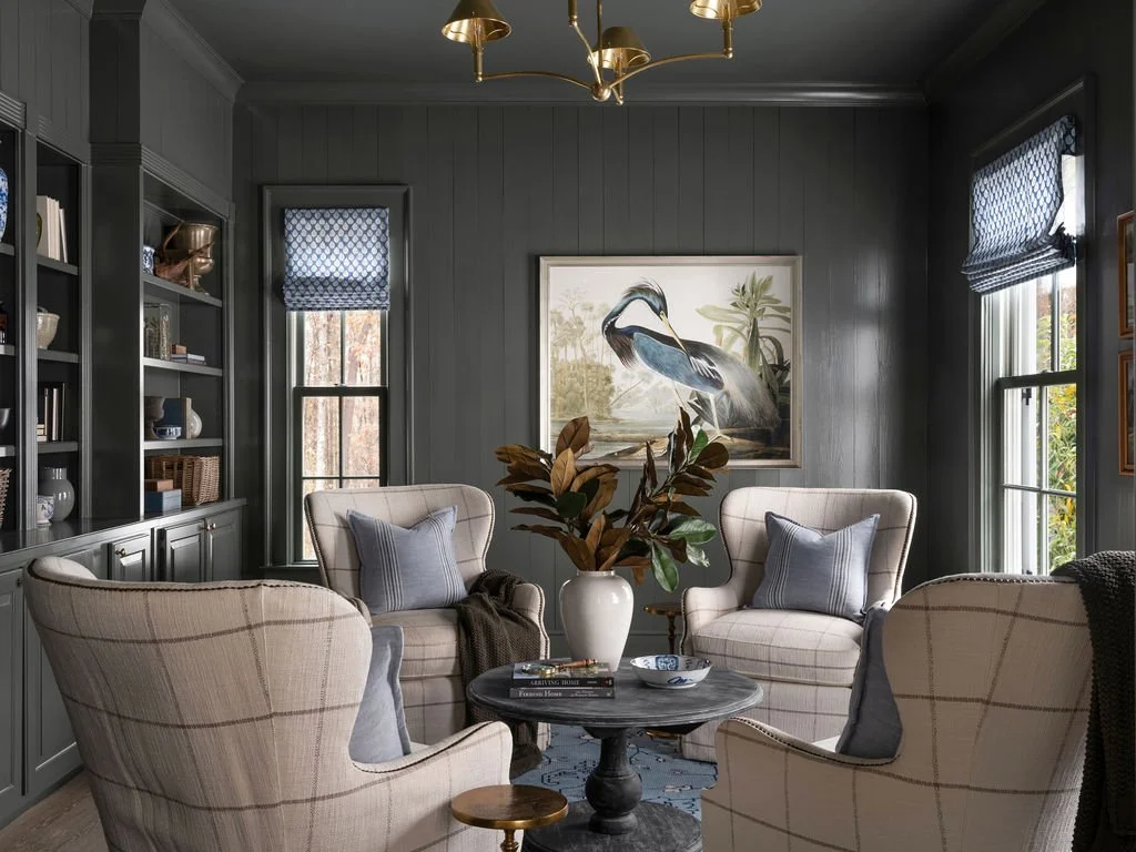 A cozy living room with grey walls, four upholstered armchairs with plaid fabric, a central round coffee table, a large plant in a white vase, and a painting of a heron on the wall. There are two windows with blue patterned curtains.