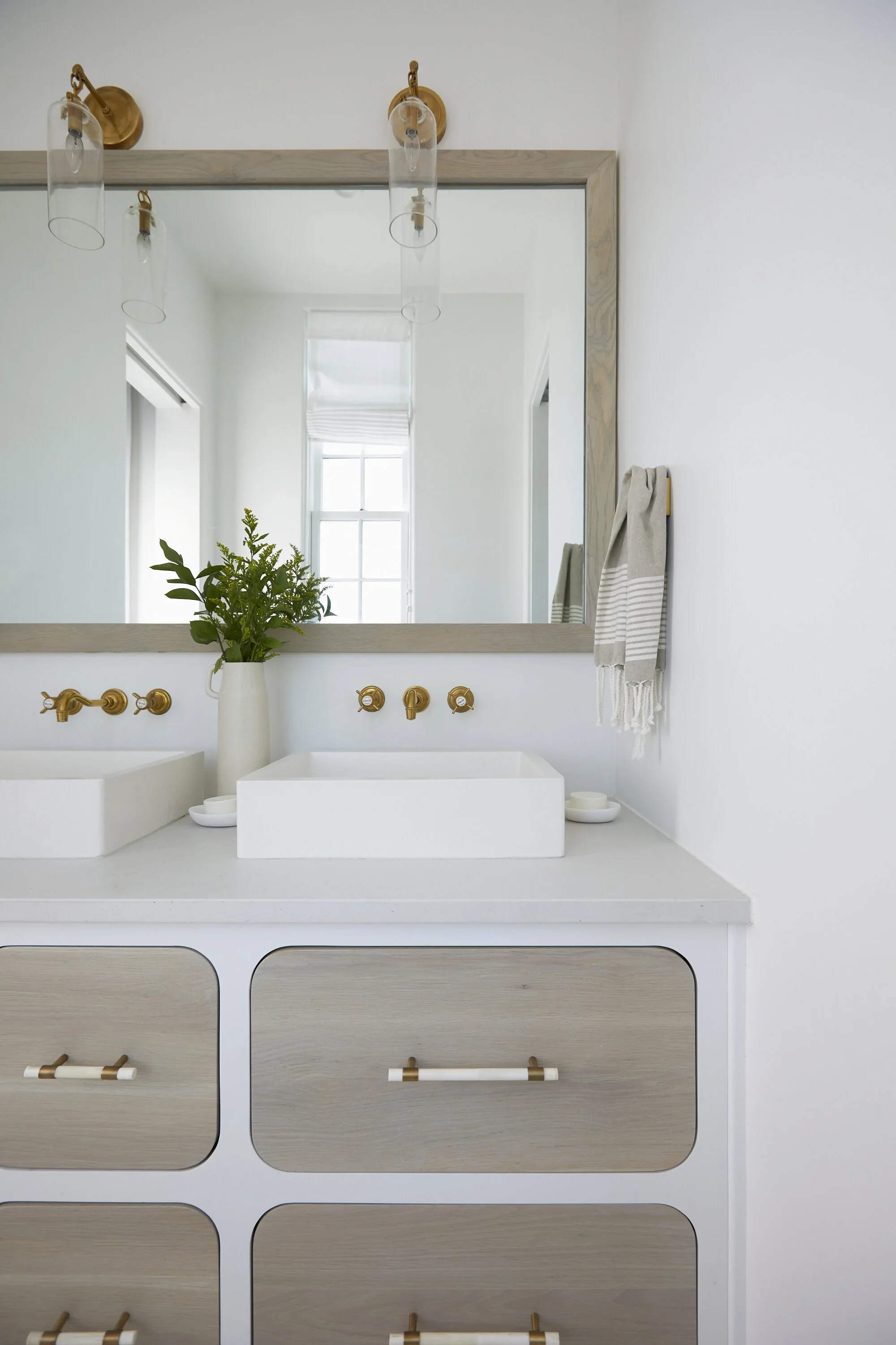 Modern bathroom vanity with a large mirror, white countertop, and rectangular vessel sinks, decorated with a vase of greenery and a gray striped towel hanging on the wall.