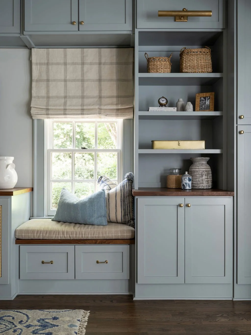 Built-in window seat with cushions and pillows beneath a window with a beige and white check Roman shade, next to a built-in cabinet and open shelves with decorative items.