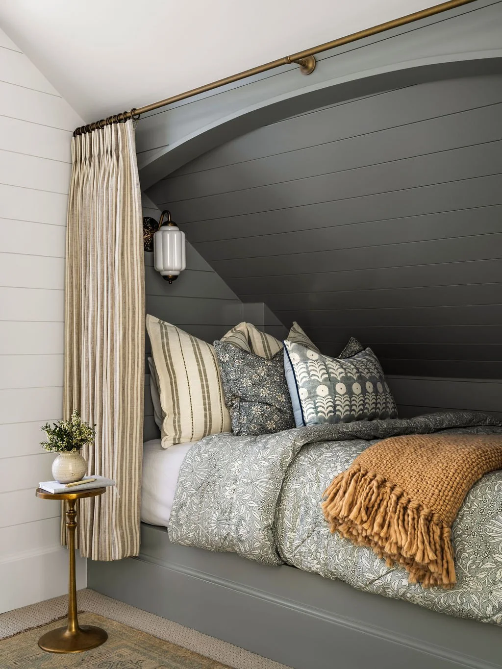 A cozy bedroom with a sloped gray ceiling, striped curtains, a small side table with a potted plant, and a bed with patterned pillows and a beige throw blanket.