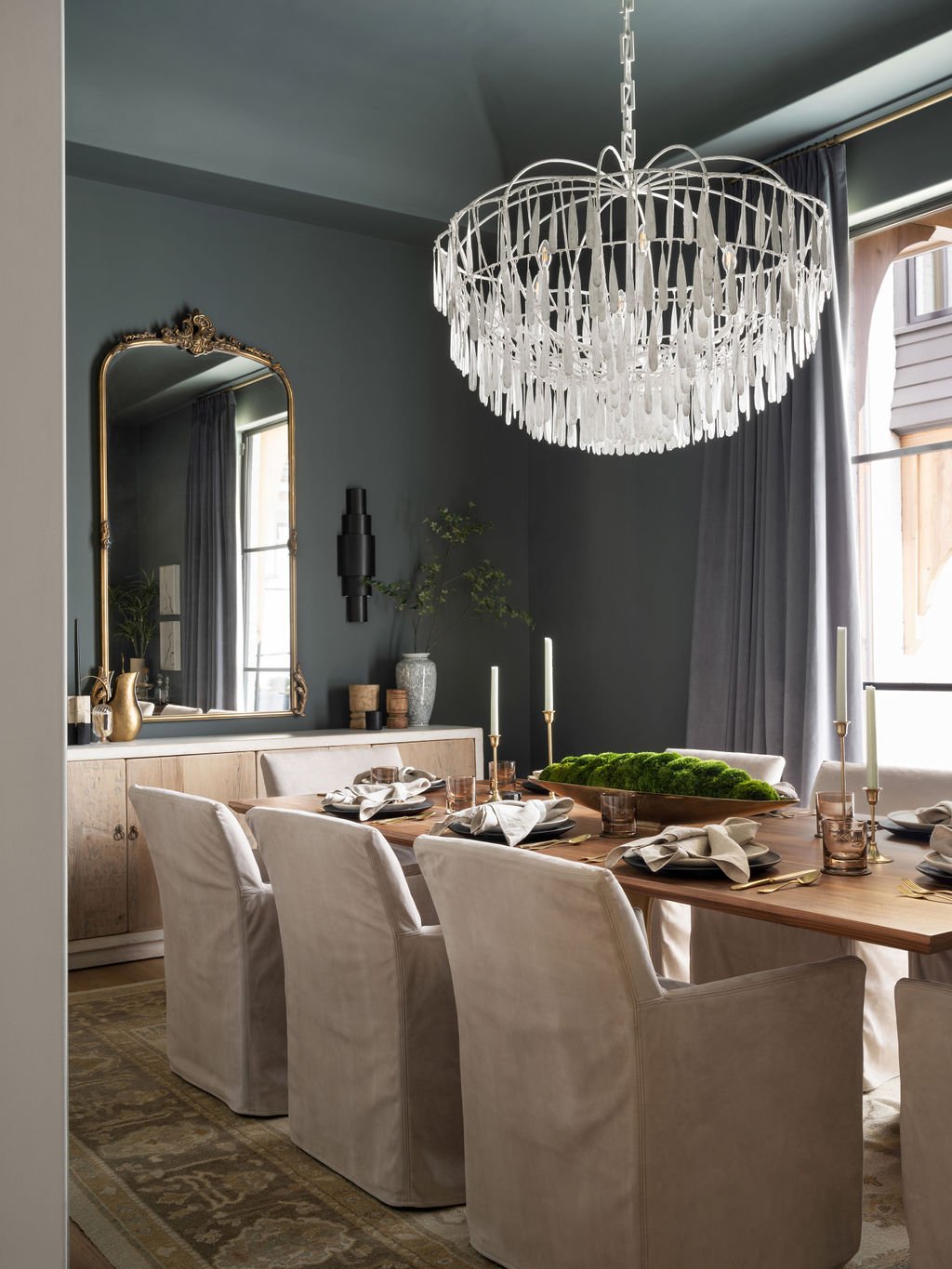 Elegant dining room with a wooden table, white upholstered chairs, and a large chandelier. The table is set with plates, napkins, candles, and a moss centerpiece. A large mirror, gray curtains, and a sideboard with decor are visible in the background