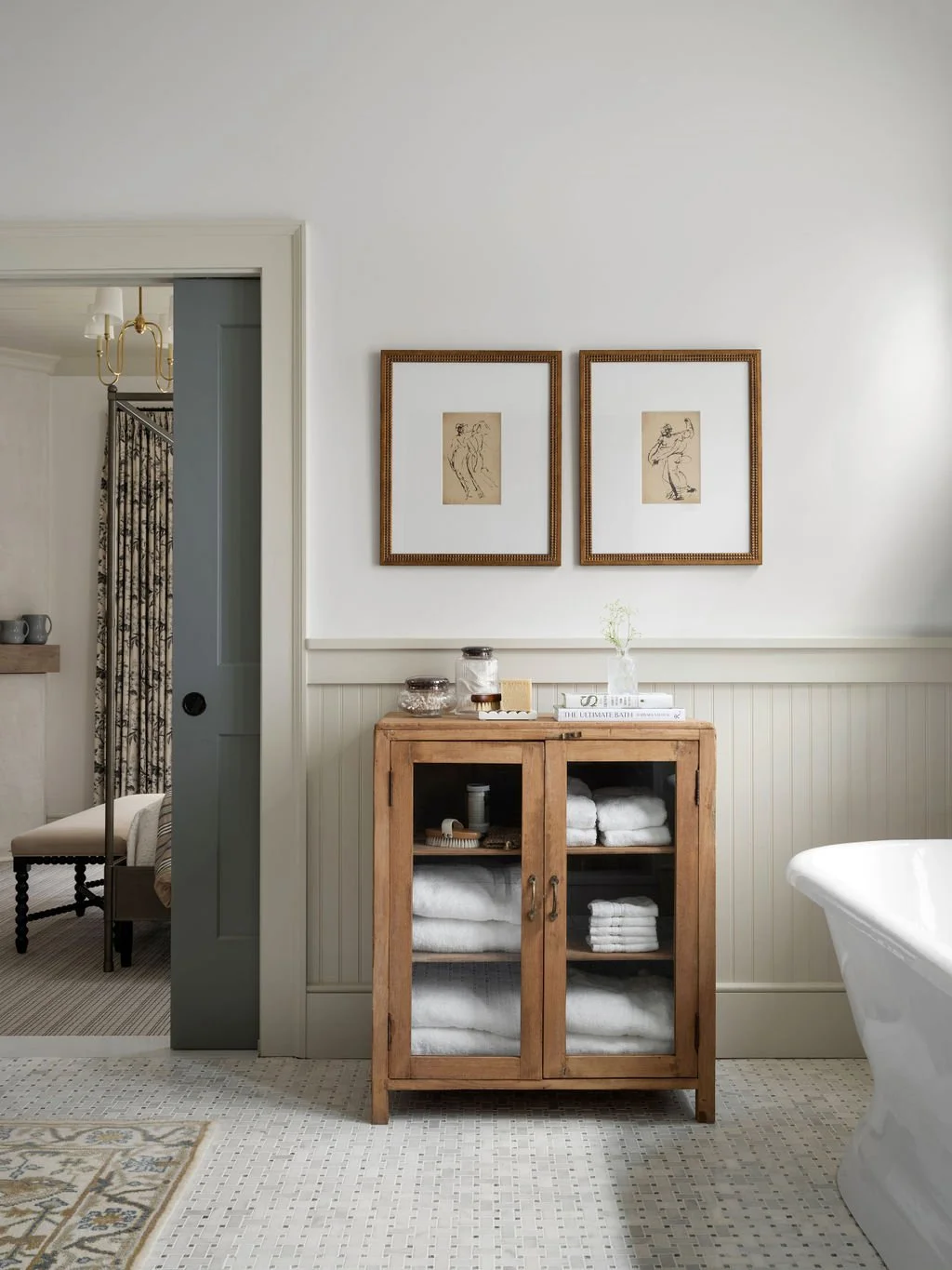 A bathroom with a wooden cabinet filled with neatly folded towels, two framed line drawings on the wall, a bathtub on the right, and a doorway leading to a bedroom with a chandelier.
