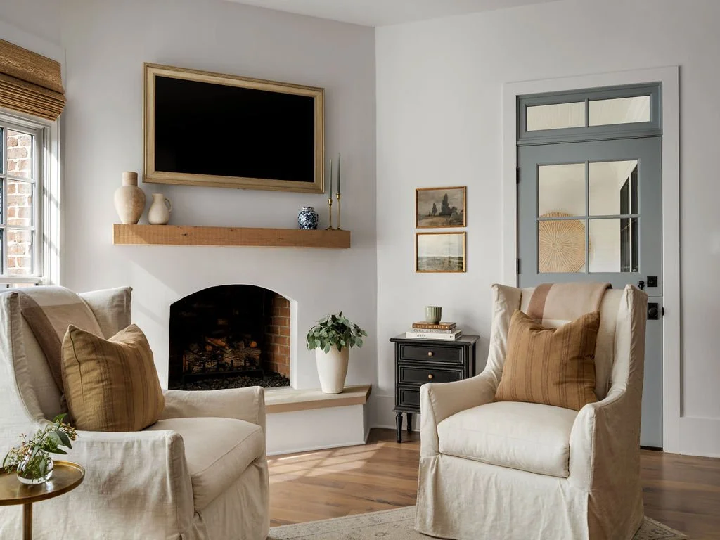 Living room with two cream armchairs and brown pillows, a small black side table, a potted plant, fireplace, wall-mounted TV, and minimalist decor.