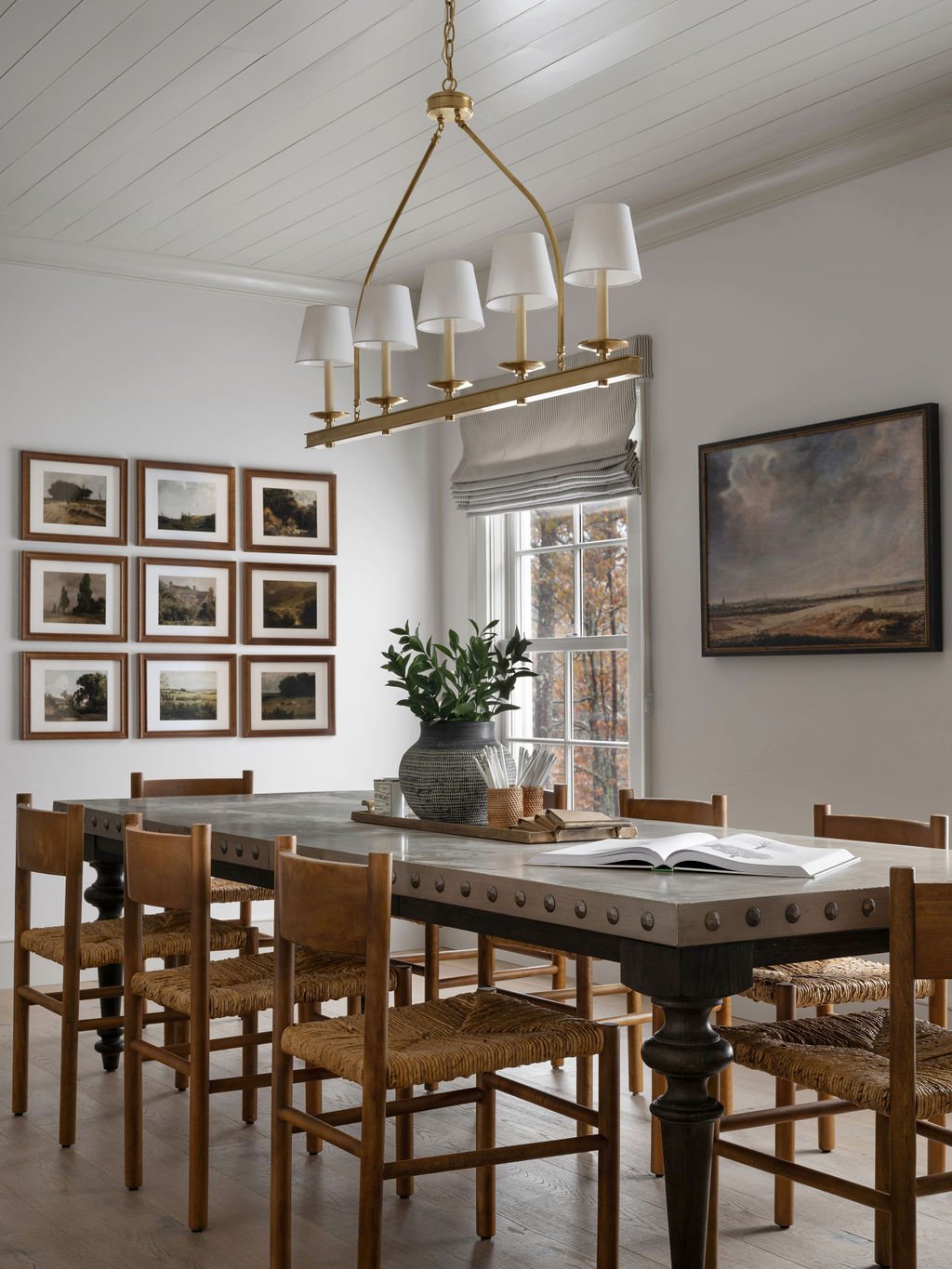 A dining room with a wooden table, eleven chairs with woven seats, a large pottery vase with plants, a set of glasses, and an open book on the table. The room has white walls, a window with a gray Roman shade, framed landscape photos on the wall, and