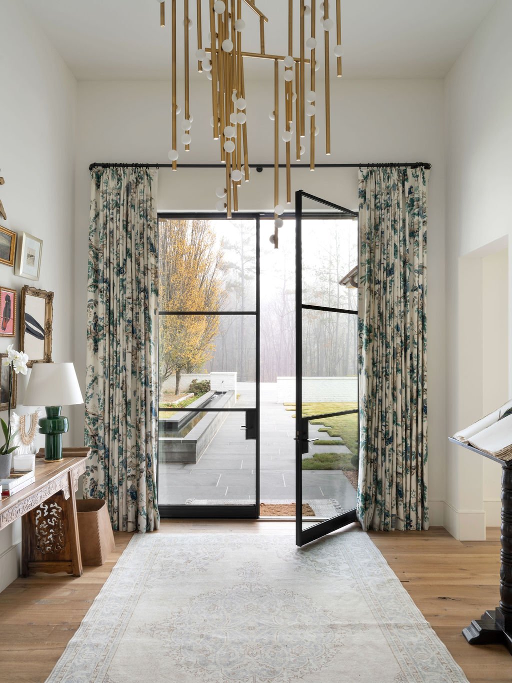 Interior view of a room with large glass sliding doors opening to an outdoor patio and garden, floral curtains, a wooden console table with a lamp, artwork on the wall, a white rug, and a modern chandelier hanging from the ceiling.
